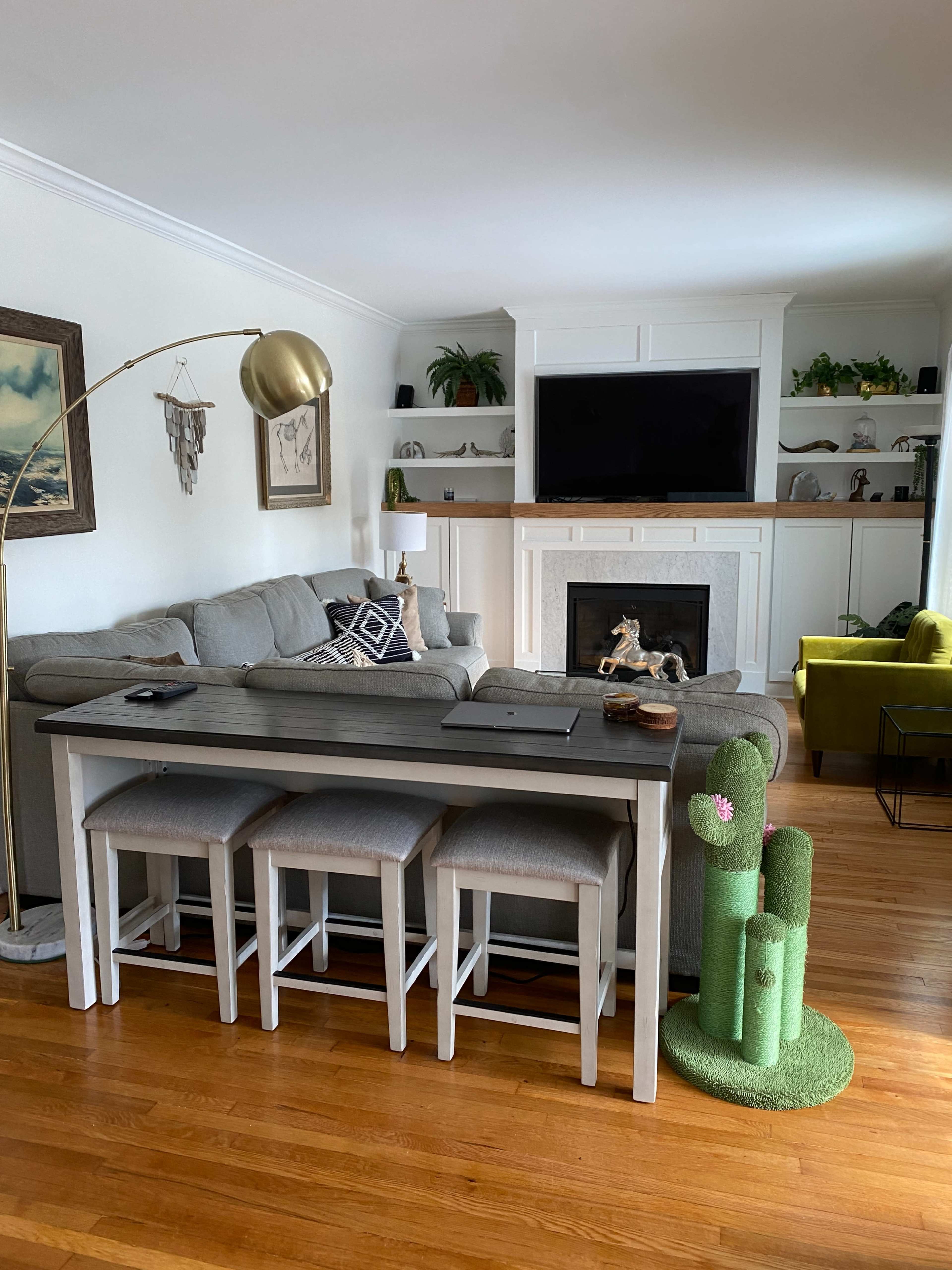 A living room with a gray sofa and a long table with three stools, set against a white fireplace and a large TV.