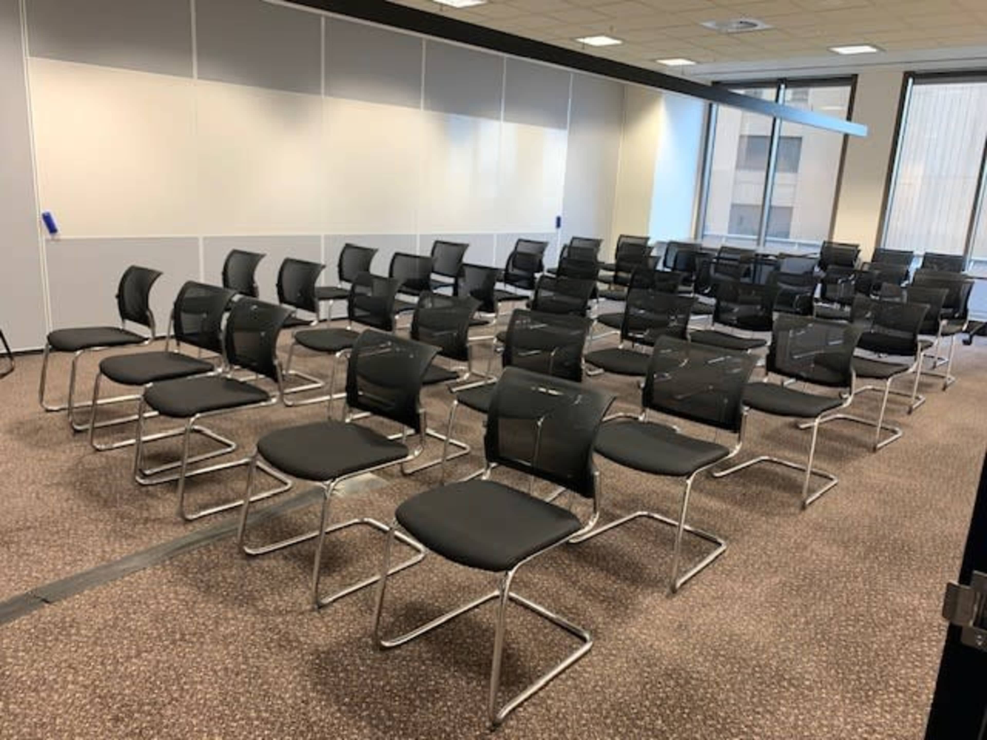 The image shows a room with rows of black metal and mesh chairs arranged in a grid pattern on a carpeted floor.