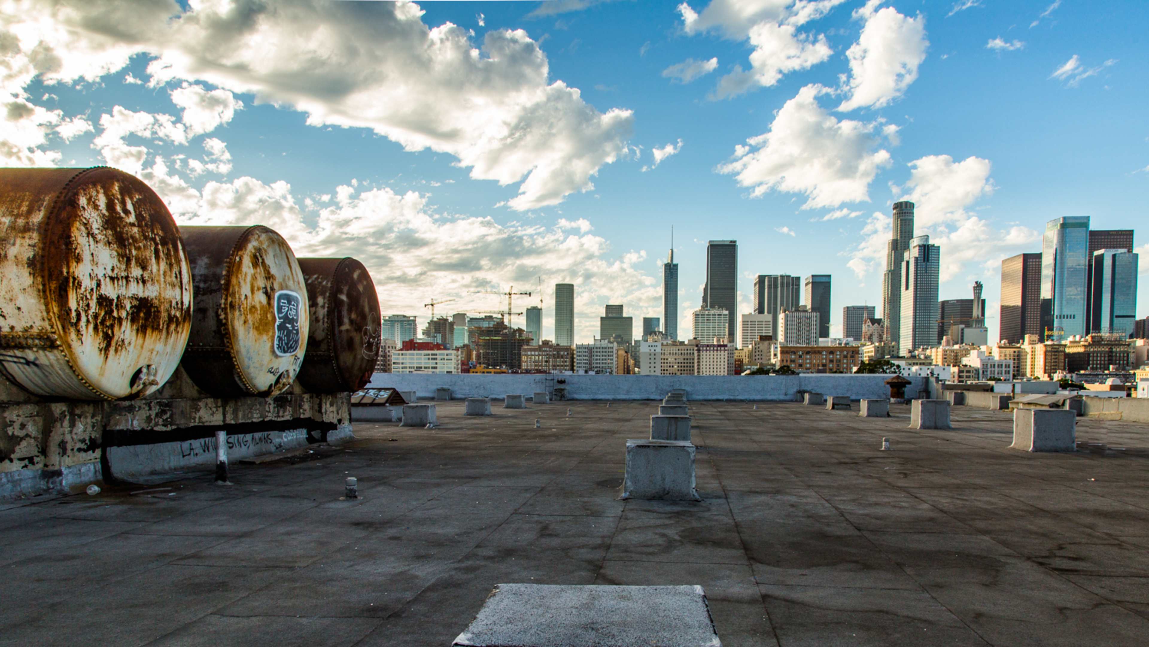 A rooftop with three large, rusty tanks overlooking a city skyline filled with tall buildings under a partly cloudy sky.