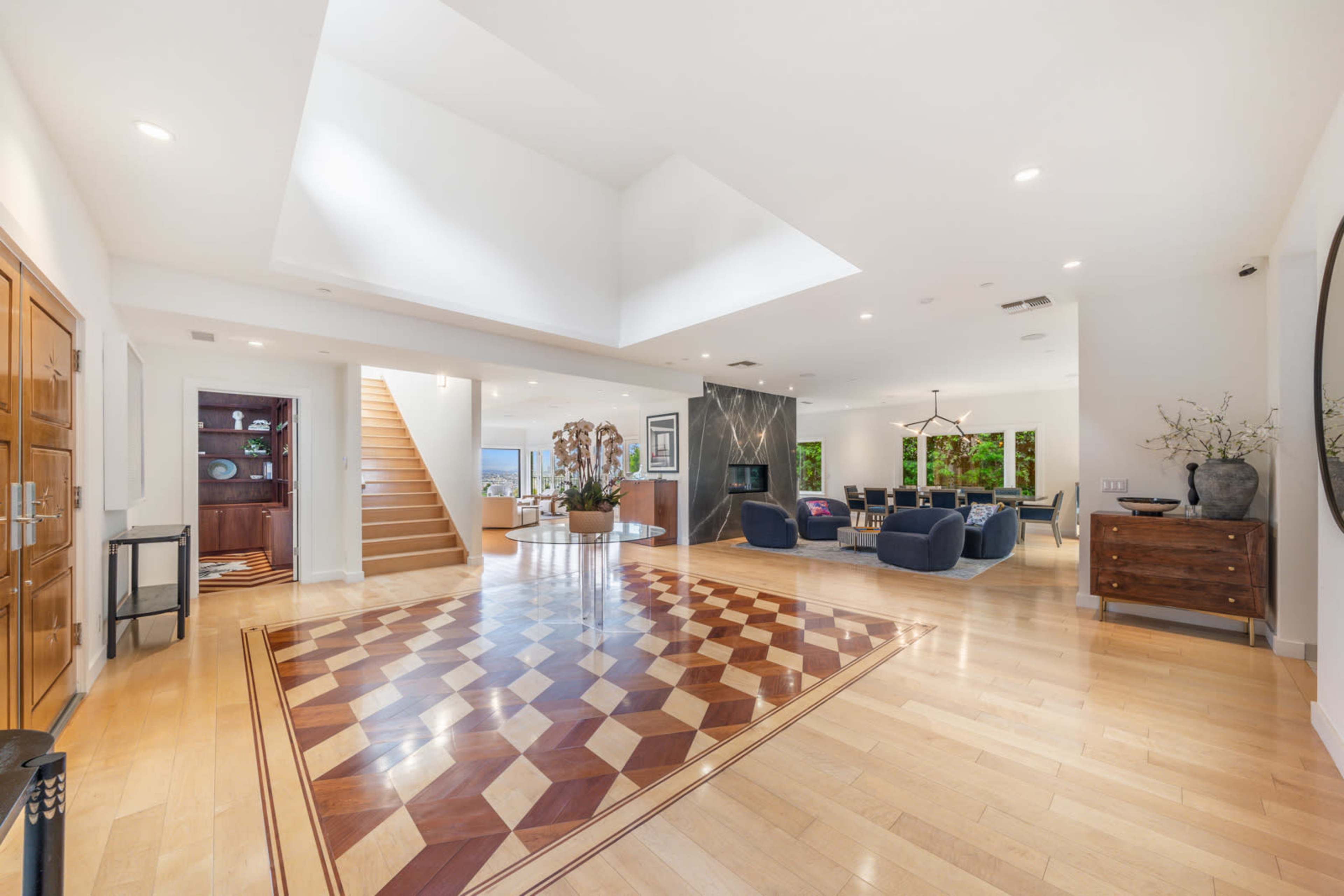 A spacious, modern living area with a patterned wooden floor, a staircase in the background, and large windows providing natural light.