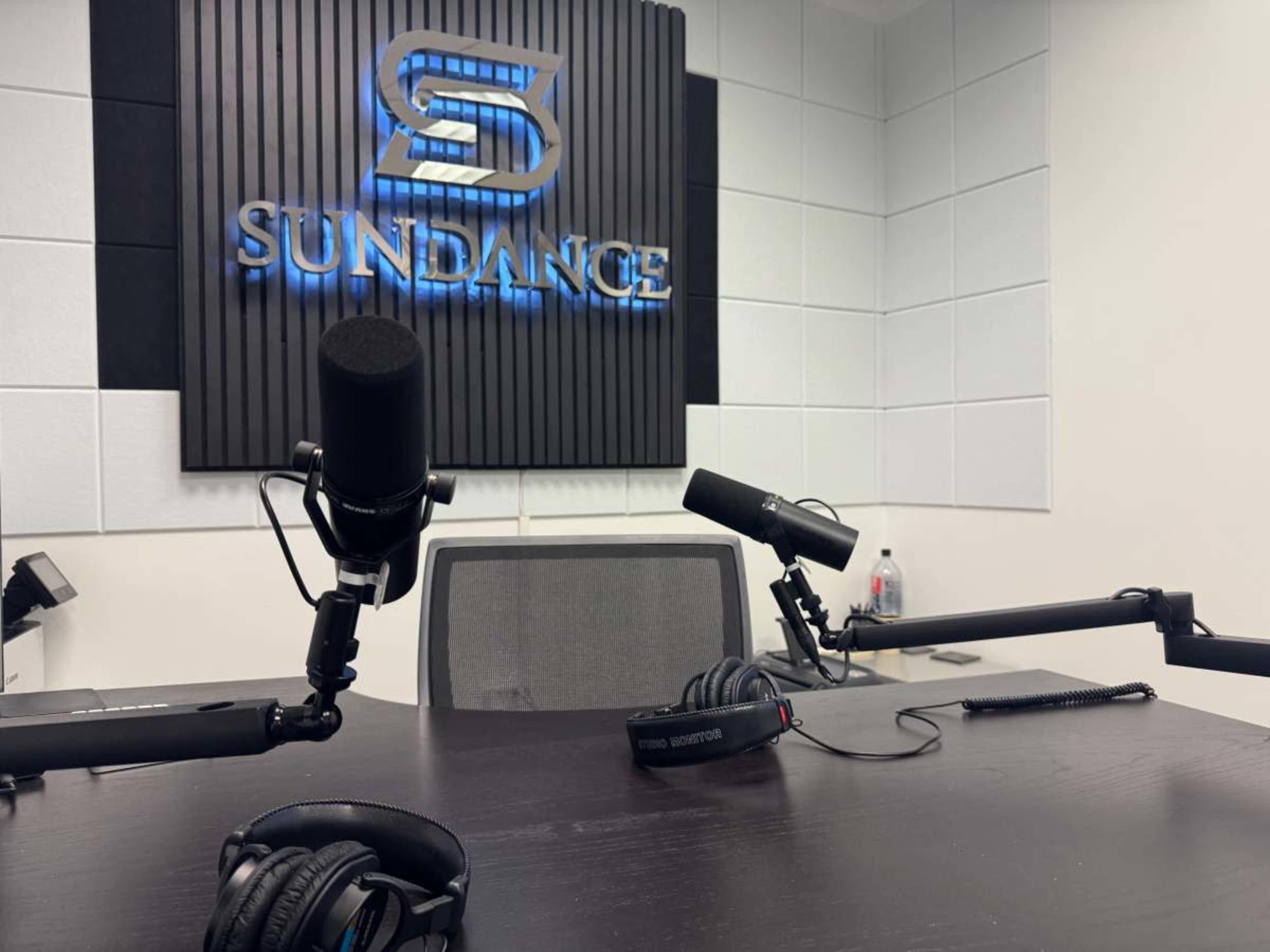 The image shows a recording studio with a large "SUNDANCE" sign on the wall, two microphones on adjustable arms, and headphones placed on the table.