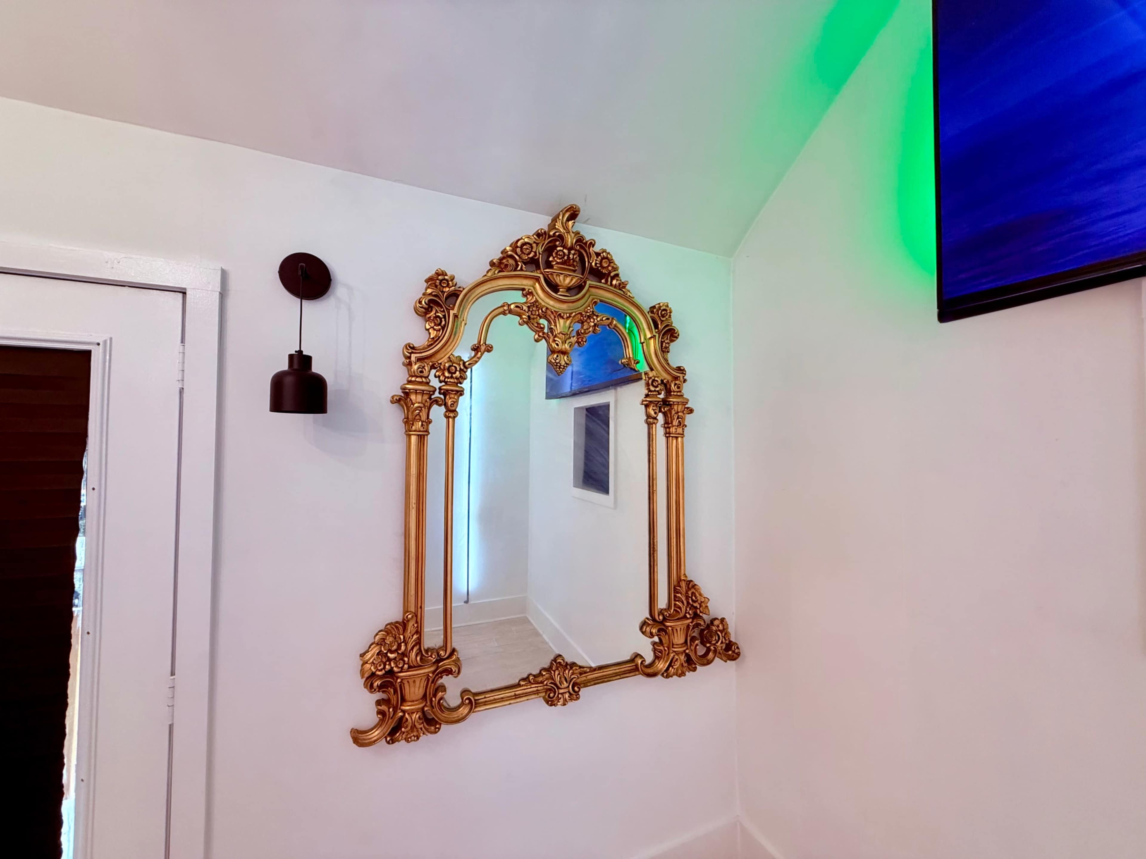 A large, ornate gold-framed mirror hangs on a white wall in a well-lit room, accompanied by modern art on the adjacent wall.