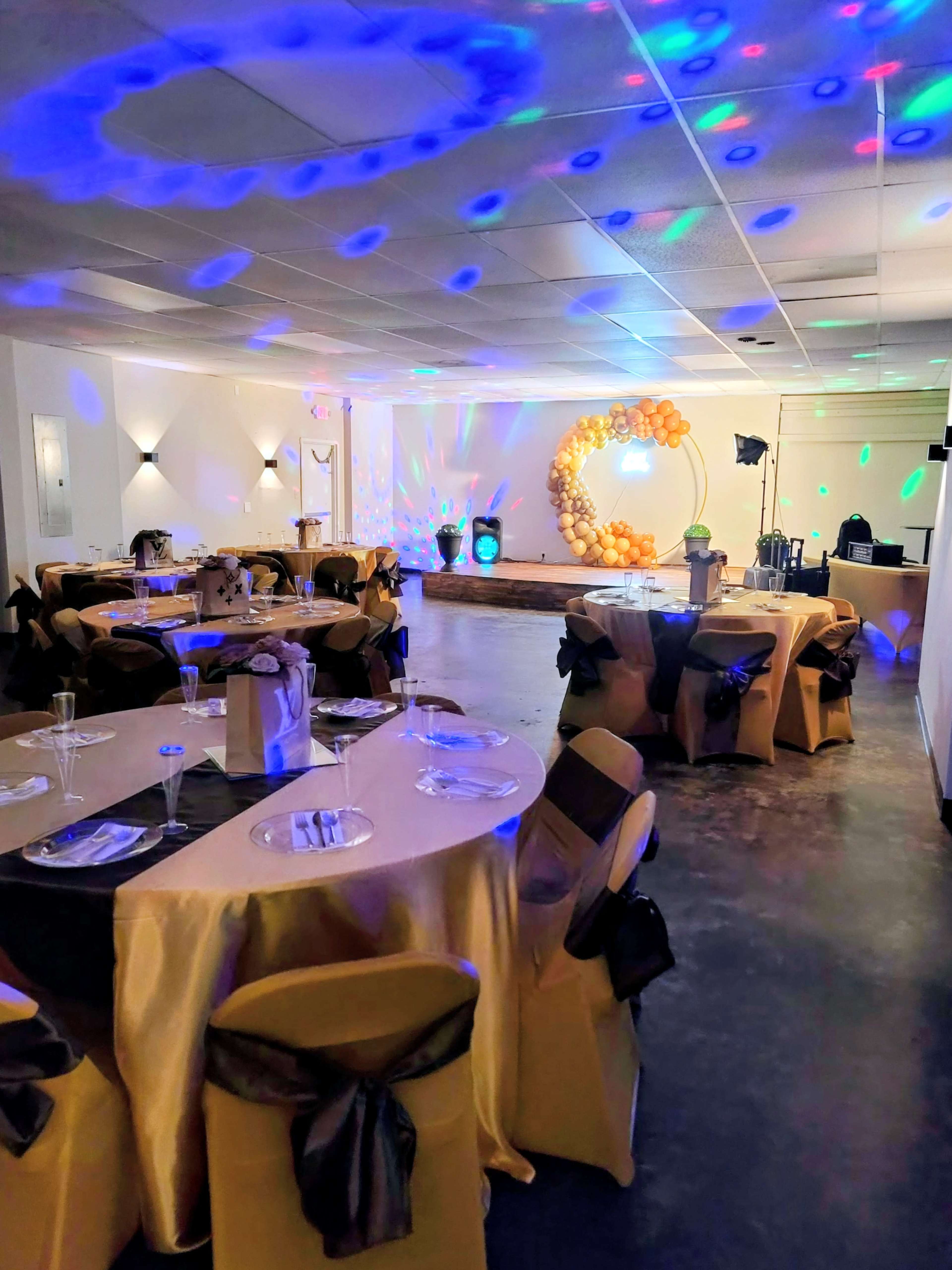 The image shows a decorated event space with round tables draped in gold tablecloths and black sashes, illuminated by colored lights and featuring a backdrop with a large balloon arrangement.