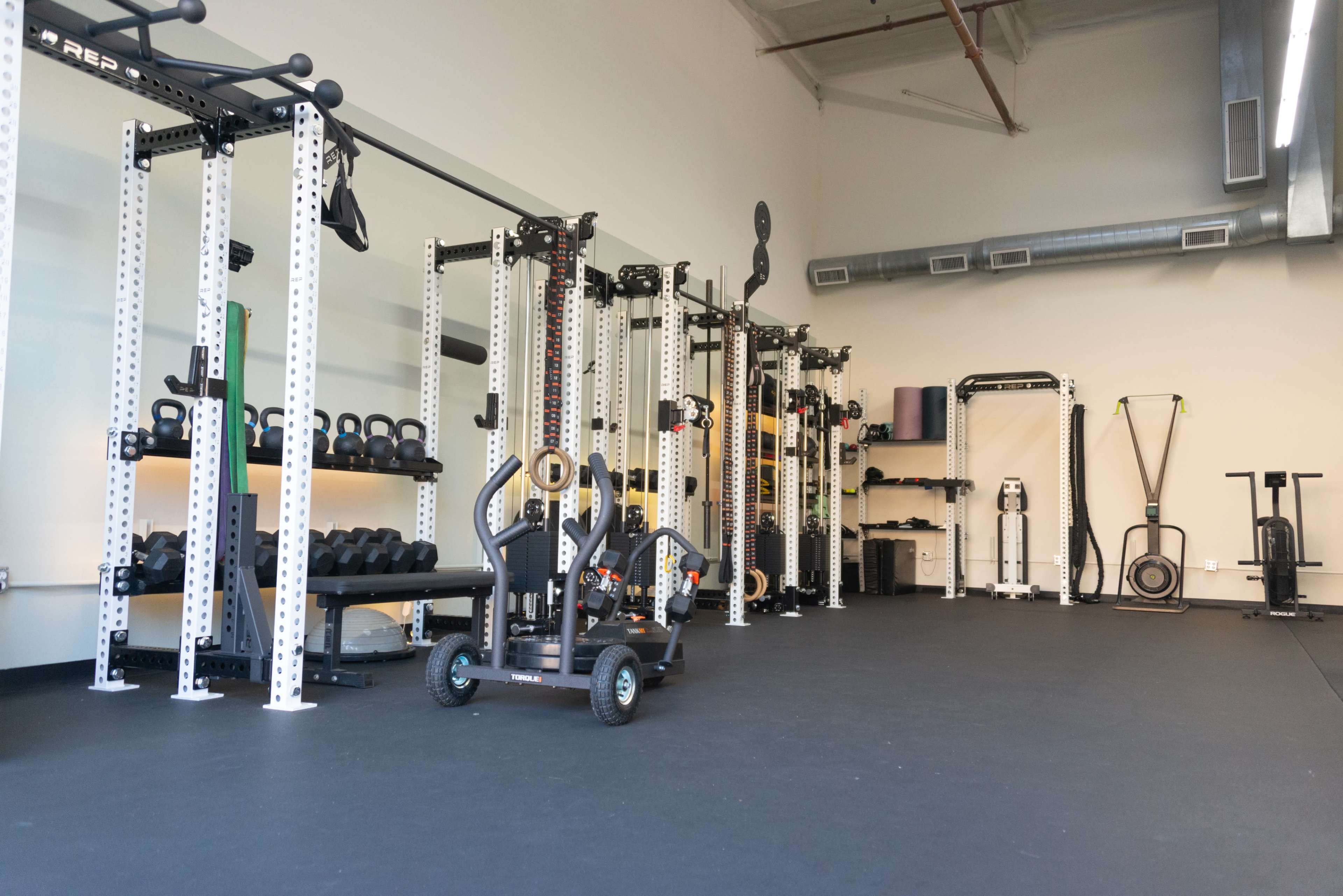 The image shows a well-equipped gym interior with various weightlifting and exercise equipment organized along the walls.