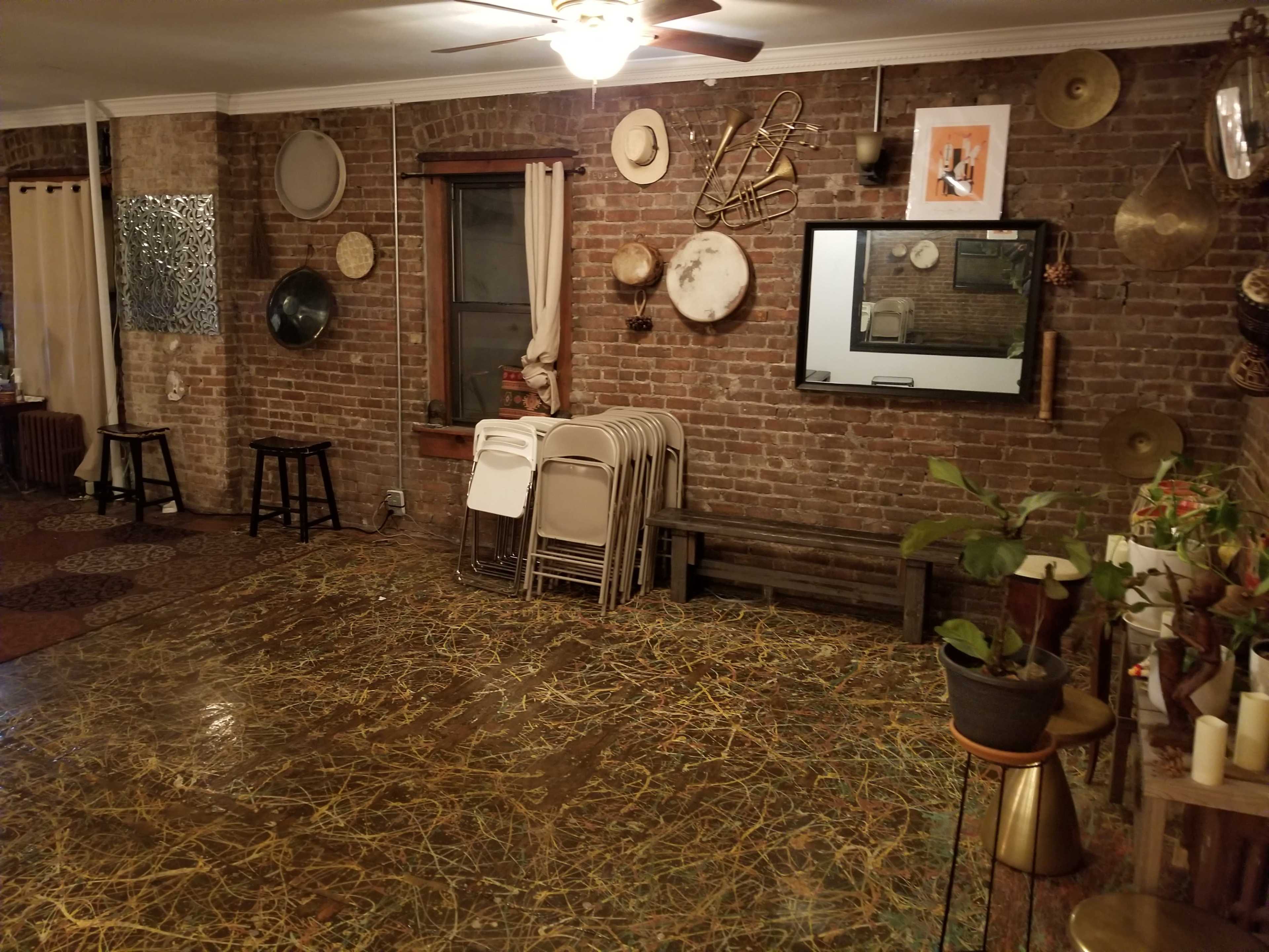 A room with exposed brick walls, featuring a mirror, various decorative items, a stack of folding chairs, and wooden furniture on a patterned floor.
