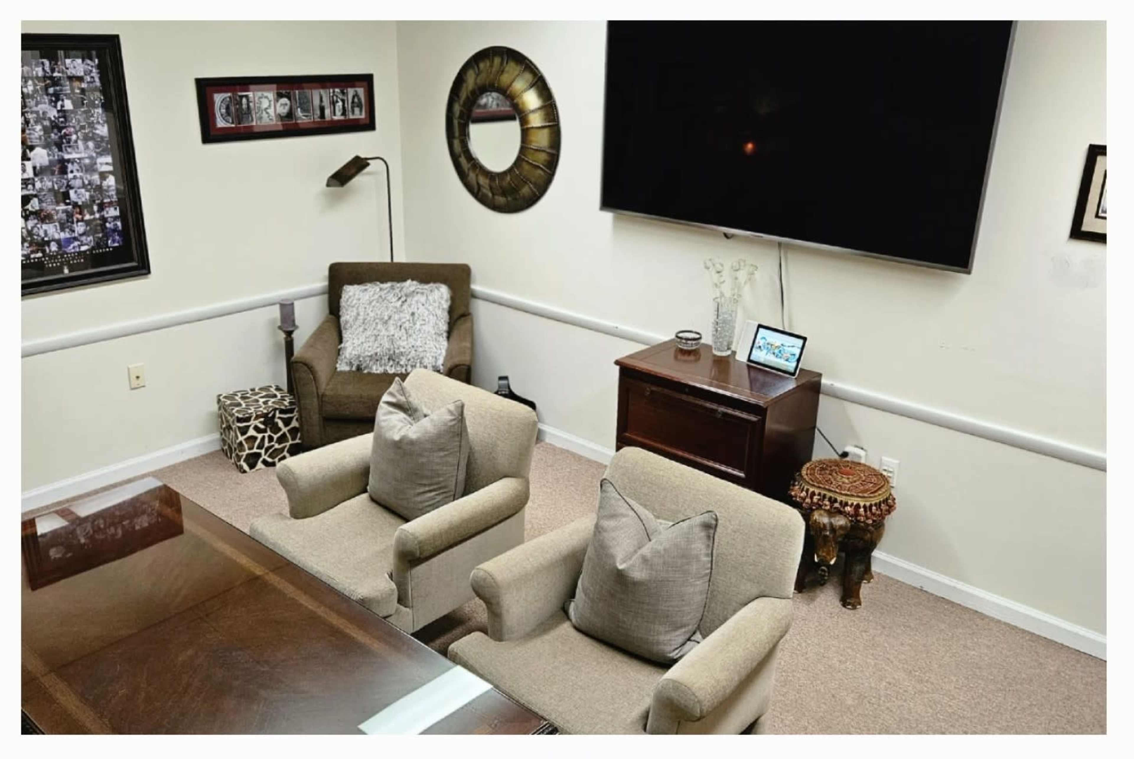 The image shows a small office space featuring two upholstered armchairs, a coffee table, a decorative mirror on the wall, and a large television mounted above a wooden cabinet.