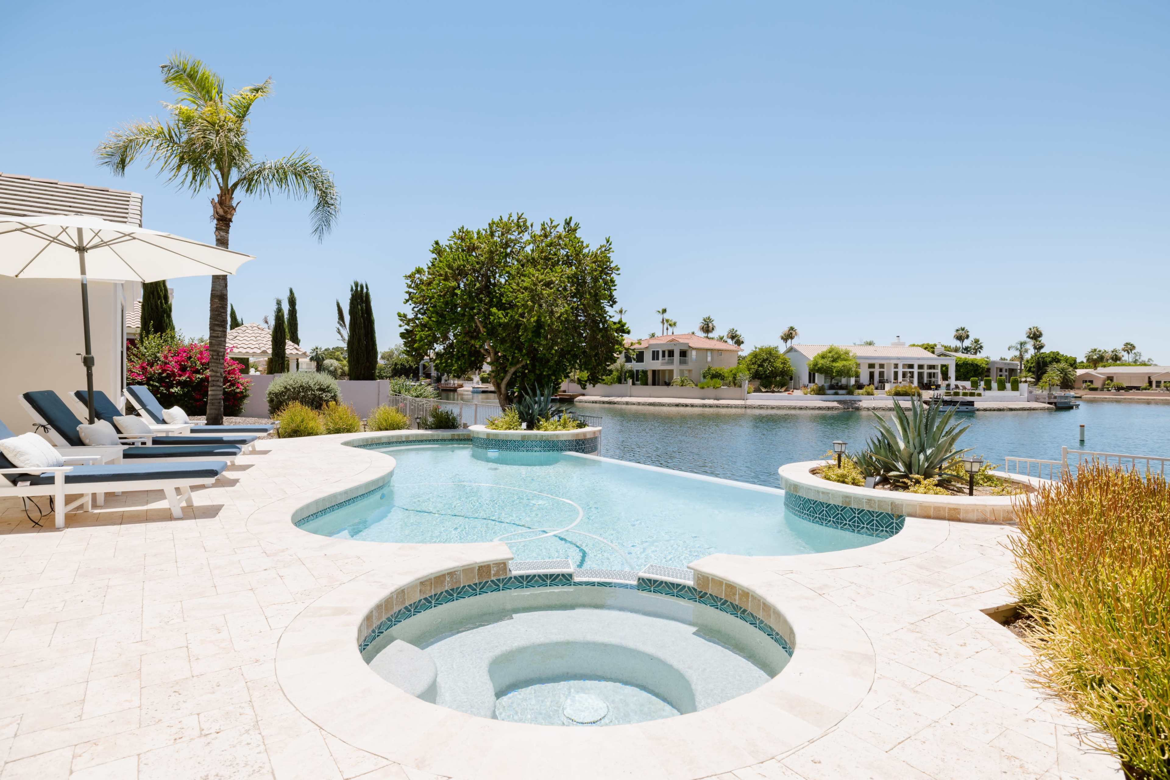 A landscaped pool area with a spa overlooking a calm body of water and palm trees in the background.
