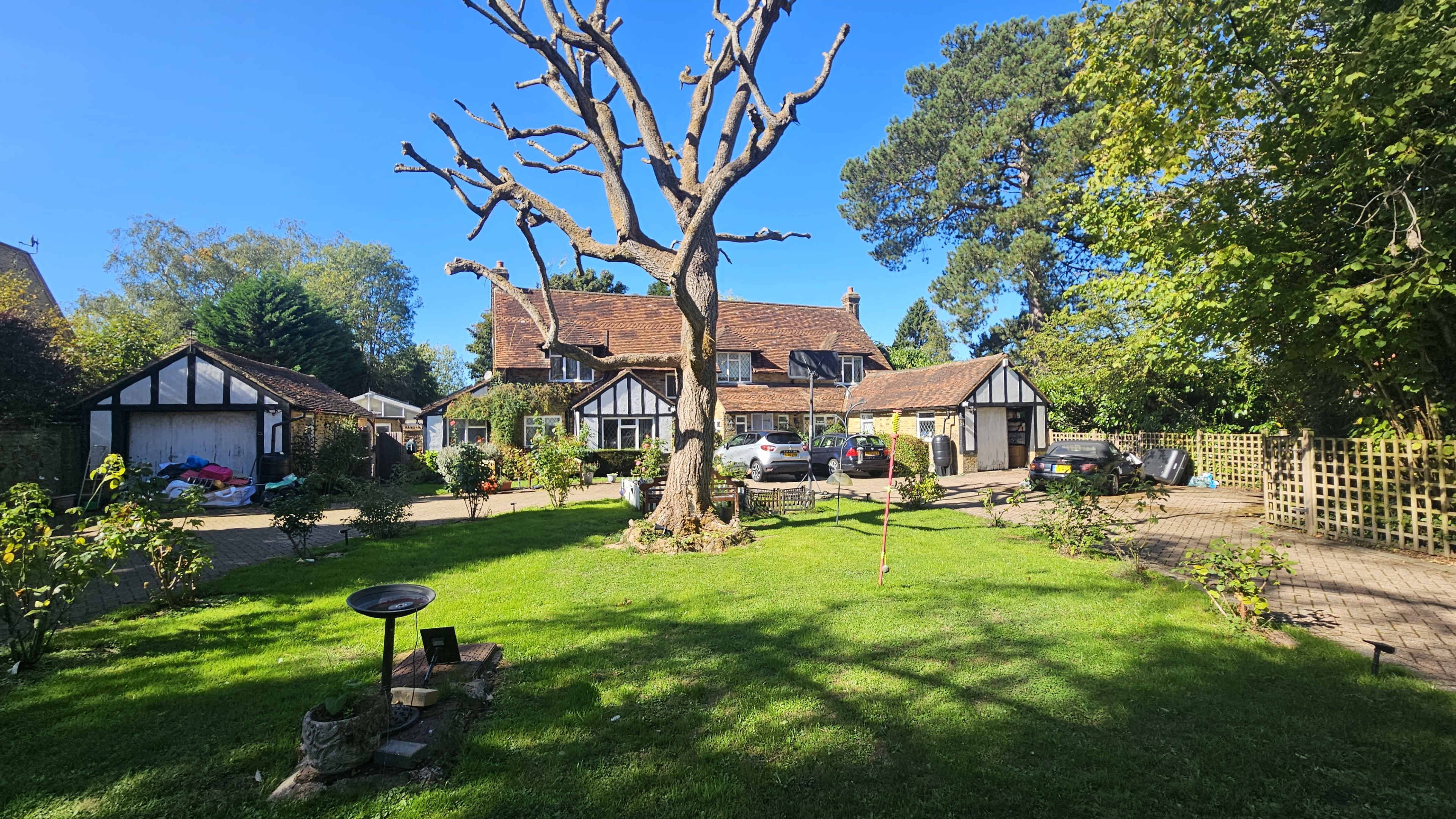 A large house with a steeply pitched roof and multiple gables is surrounded by a grassy area and a leafless tree.