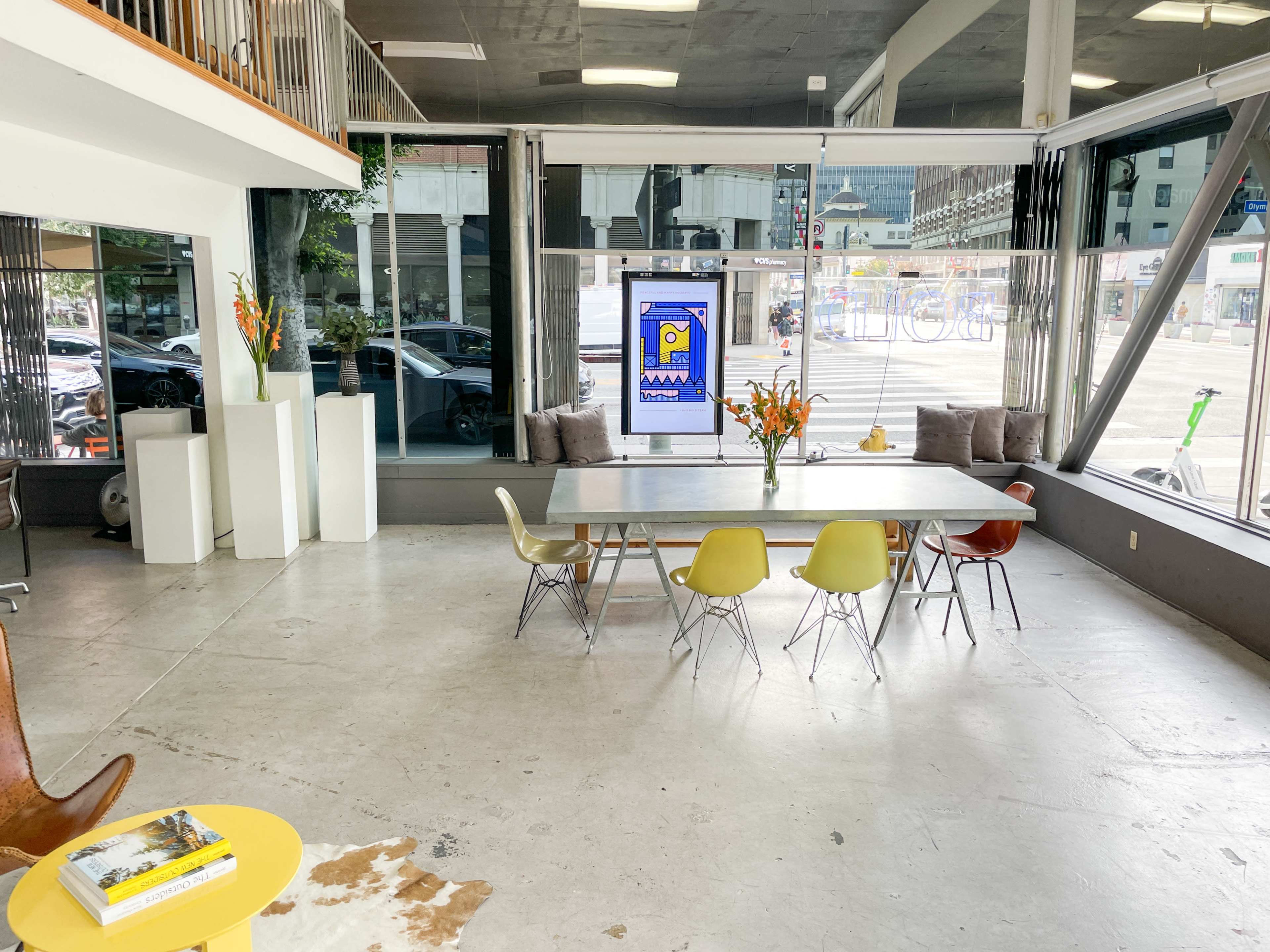 The image shows a modern, open-concept office space with a large table surrounded by colorful chairs, large windows letting in natural light, and a digital display in the background.
