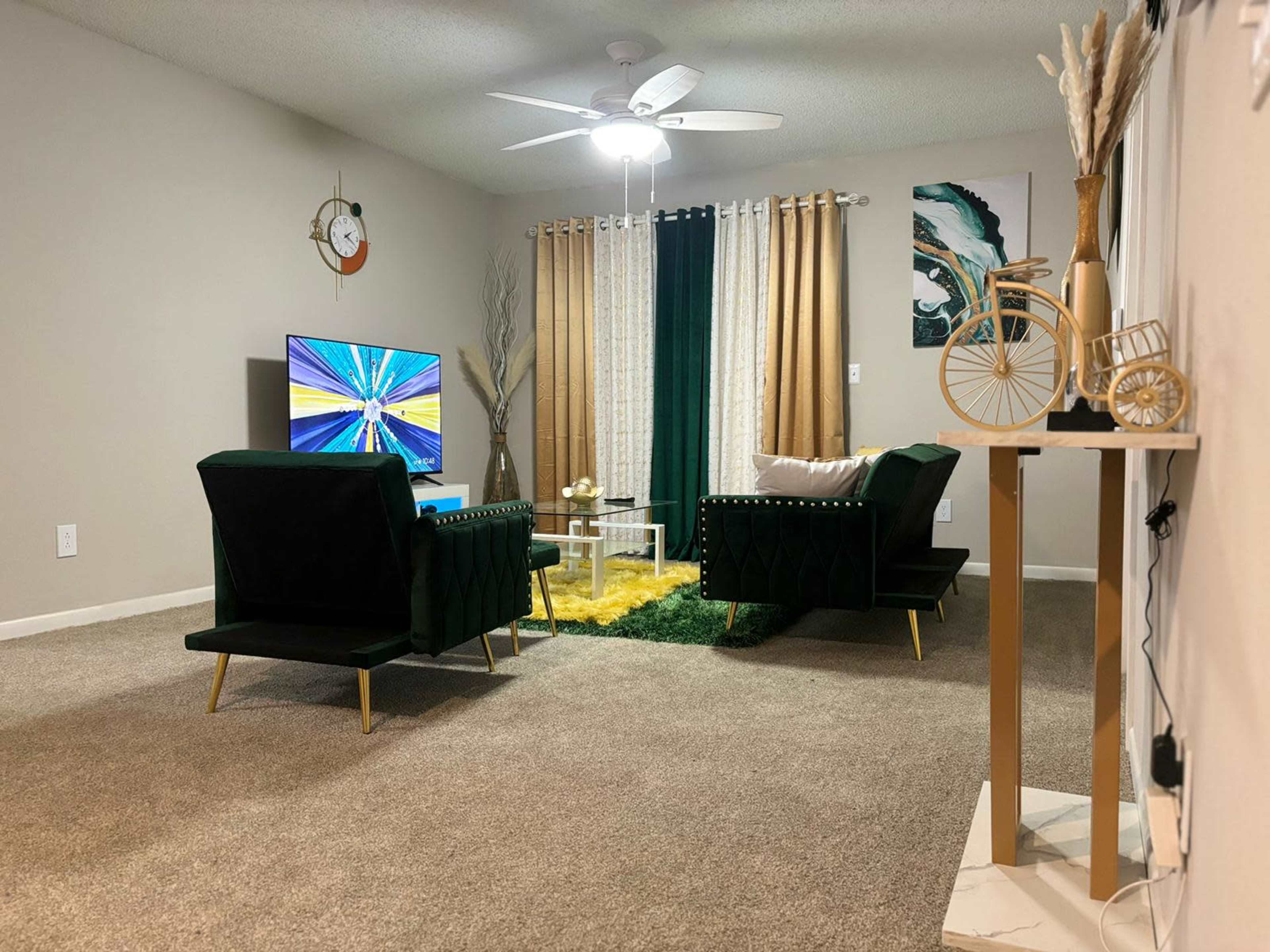 A living room features two green chairs, a television on a stand, and decorative elements, including a small table and a bicycle sculpture, with curtains and a rug adding texture to the space.