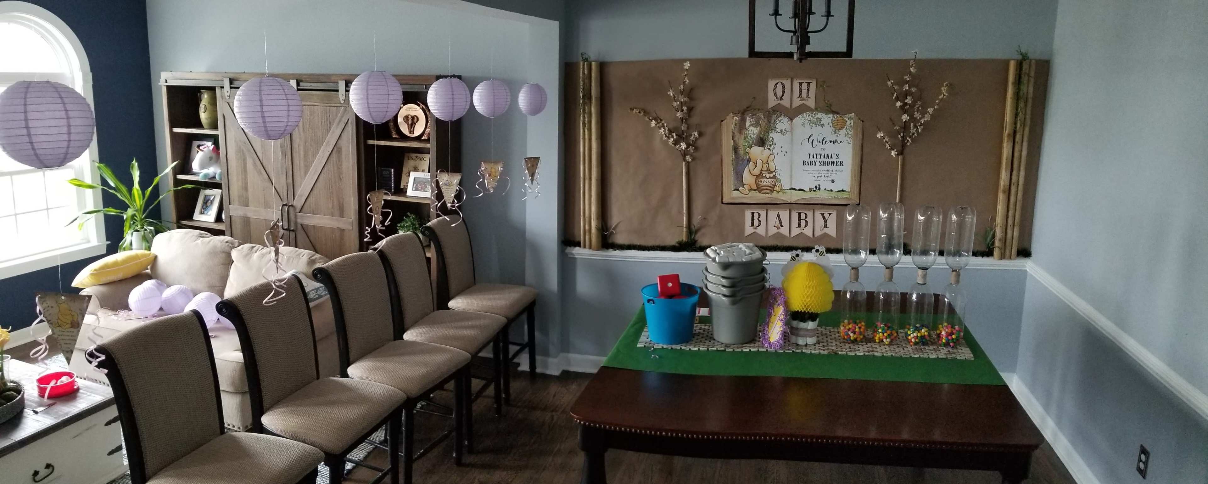 A decorated room features a table set with party supplies and a banner reading "Oh Baby," alongside a row of chairs and hanging paper lanterns.