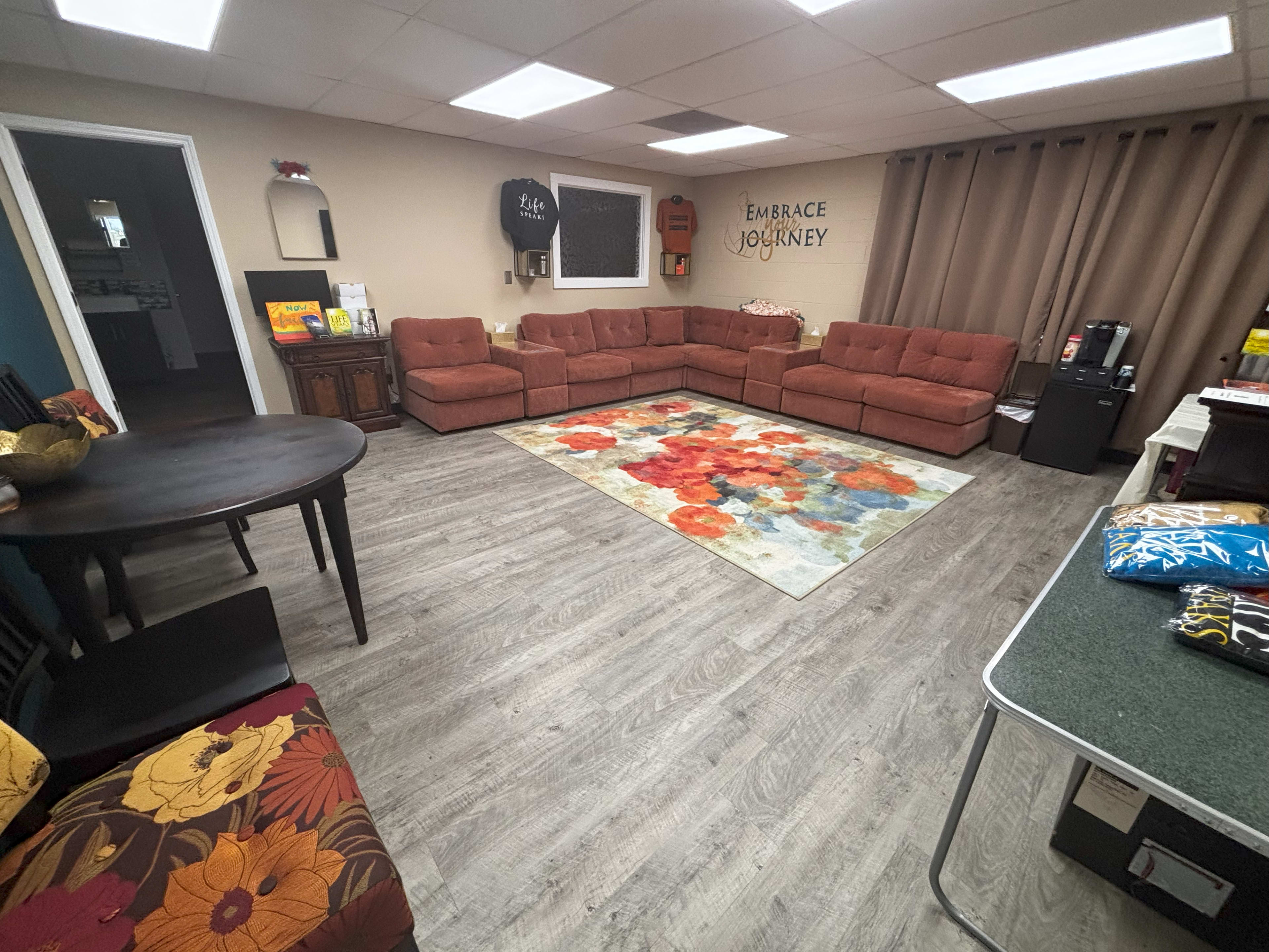 The image shows a cozy room with a circular arrangement of brown sofas, a large colorful rug, and a table set in a corner, along with various decorations on the walls.