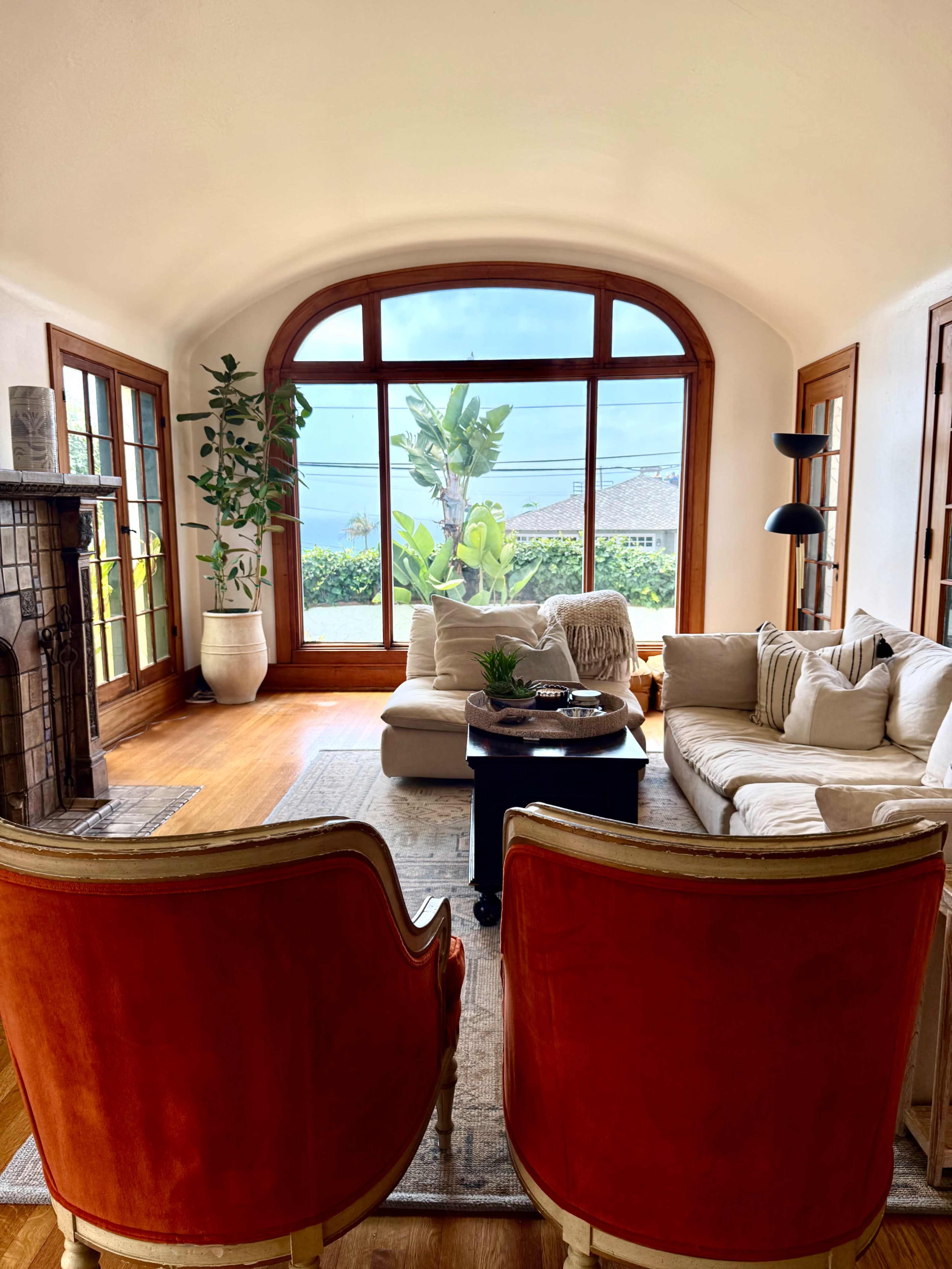 A light-filled living room features a large arched window, a beige sofa, two red upholstered chairs, and a coffee table adorned with decorative items.
