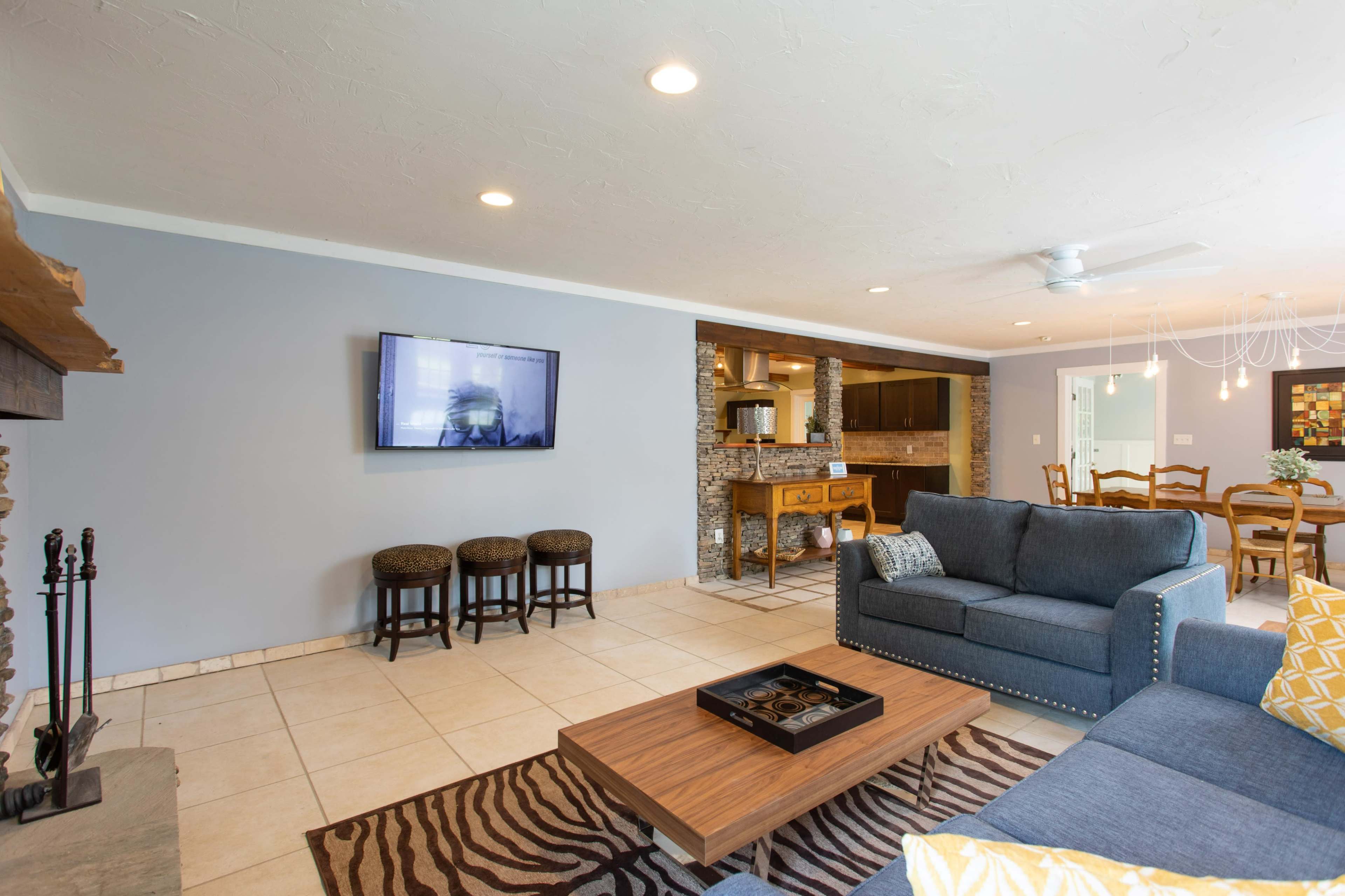 A modern living room with a blue sofa, a wooden coffee table, and a wall-mounted television, adjacent to a dining area and kitchen.