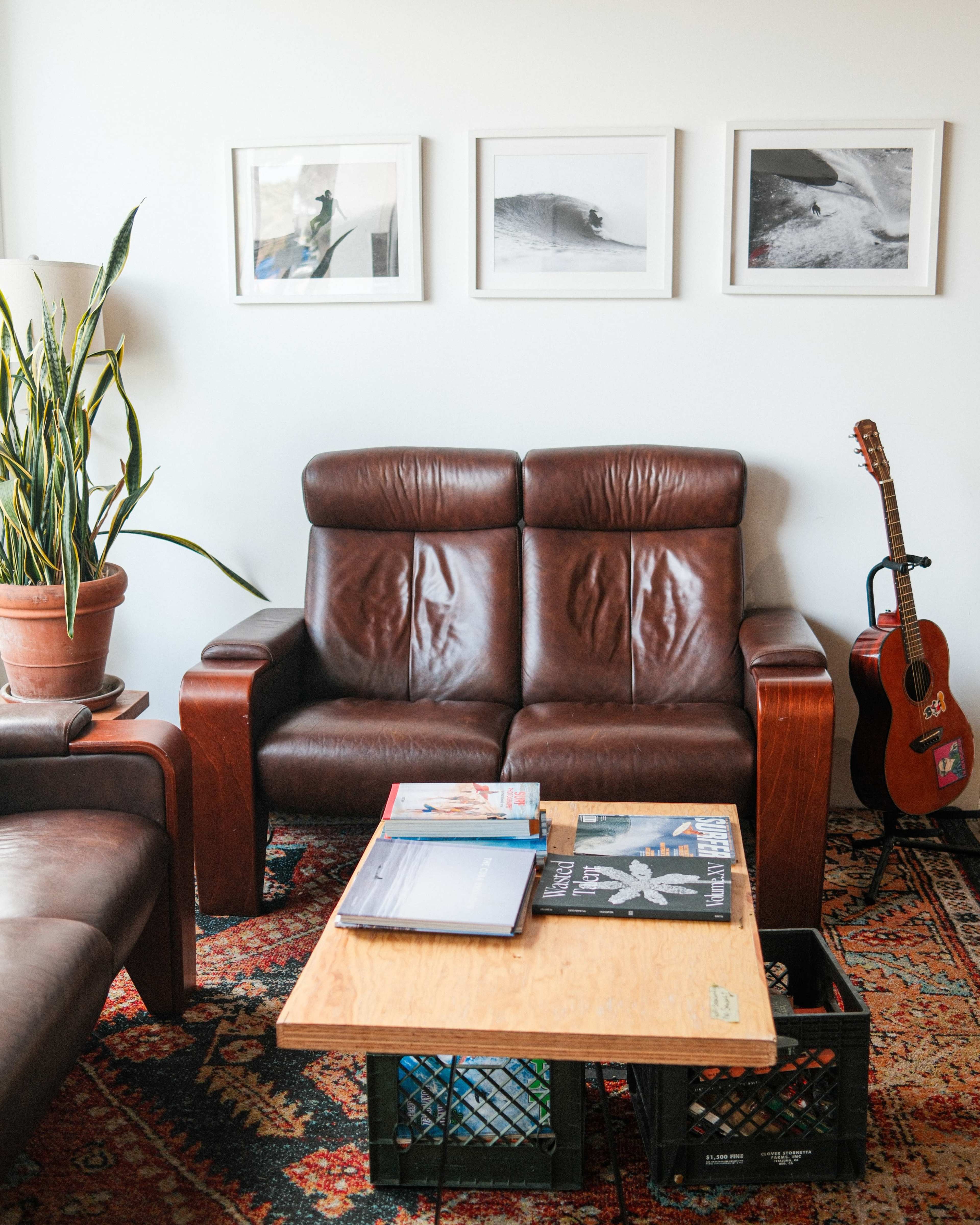 A cozy living room features a brown leather loveseat, a wooden coffee table stacked with magazines, and a nearby guitar resting against the wall alongside potted plants and framed photographs.