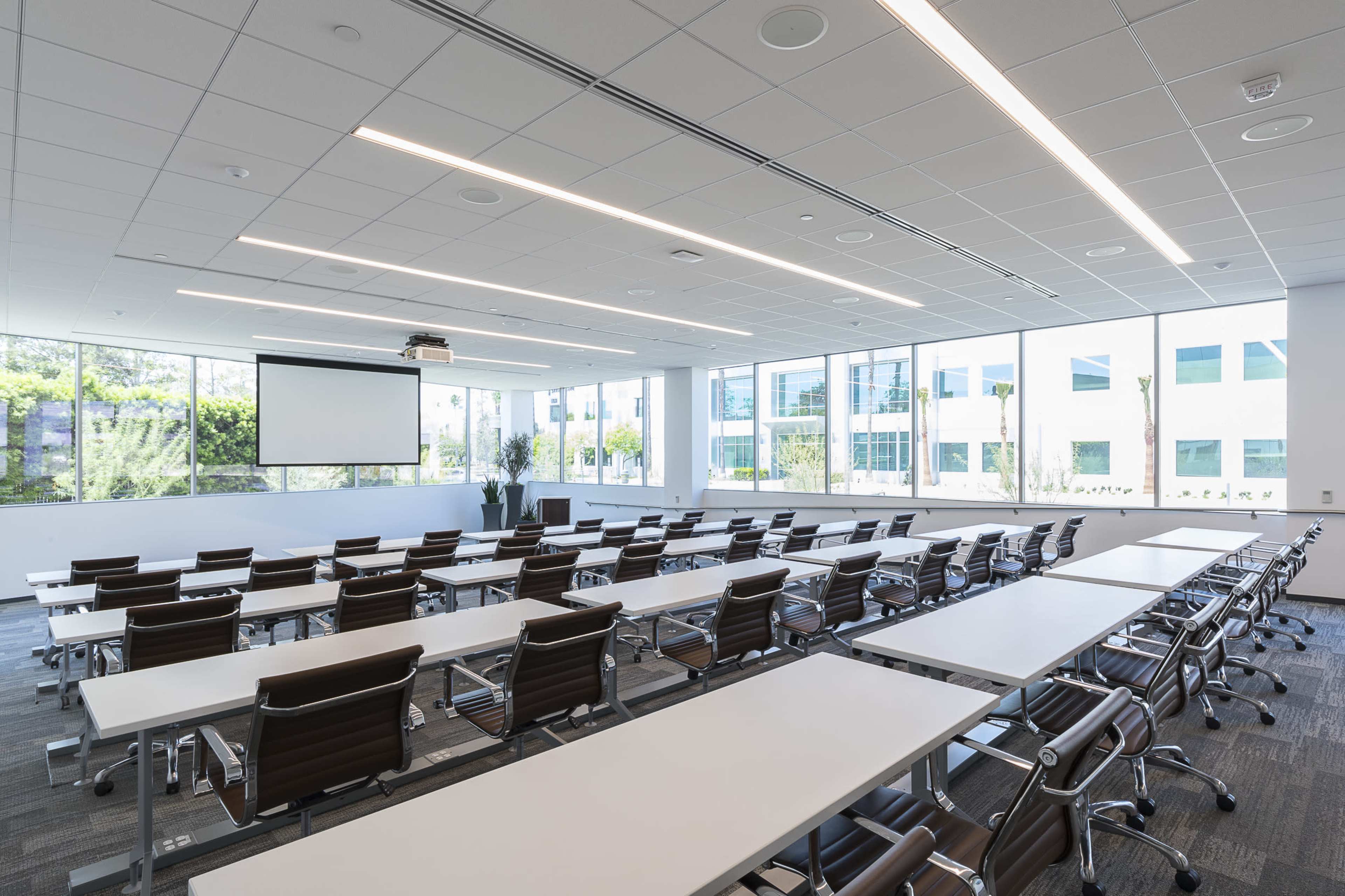 A modern conference room features rows of tables and chairs arranged toward a projection screen at the front, with large windows providing natural light.