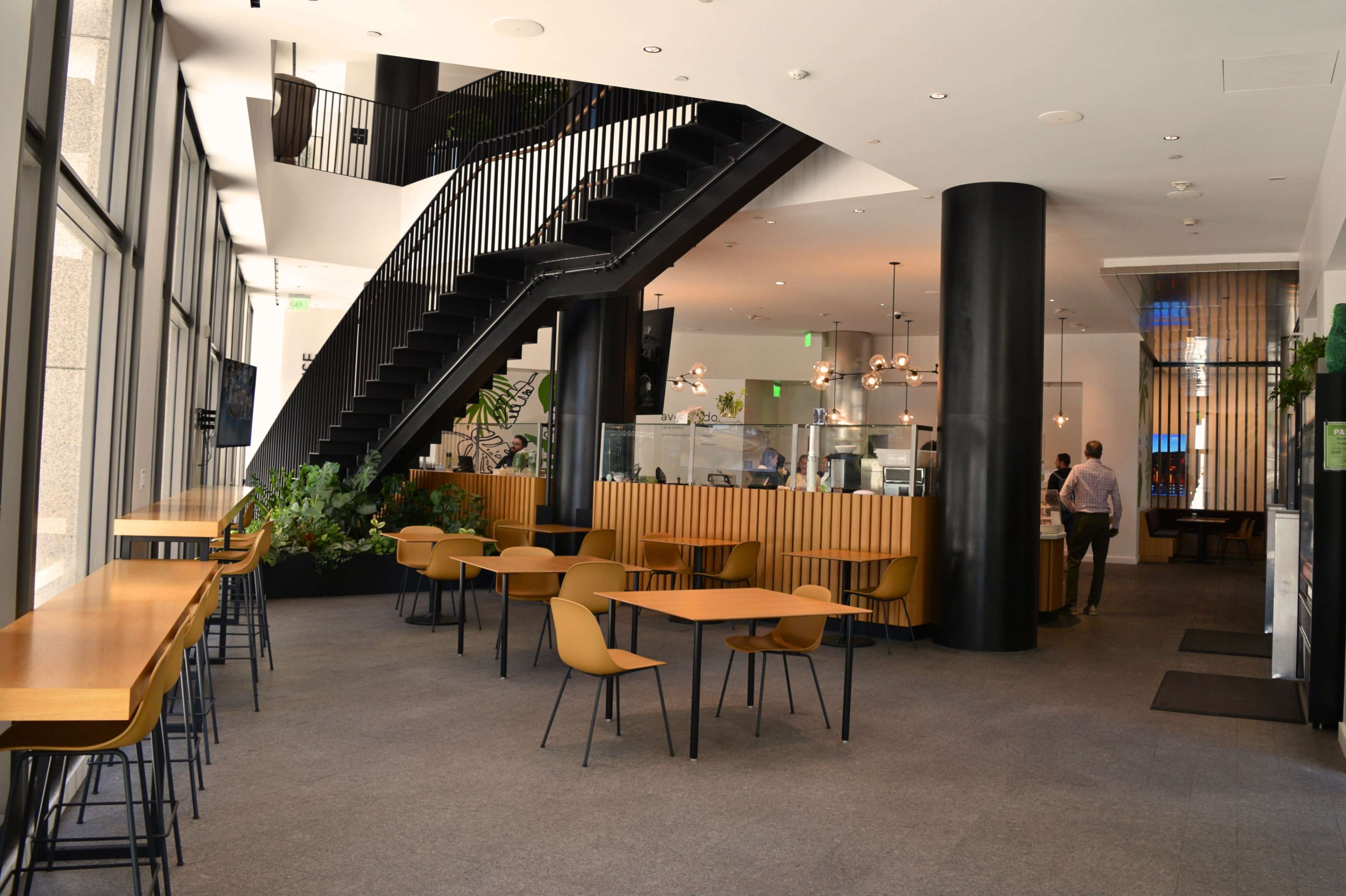 The image shows a modern interior of a café with a staircase, seating areas with tables and chairs, and a service counter in the background.