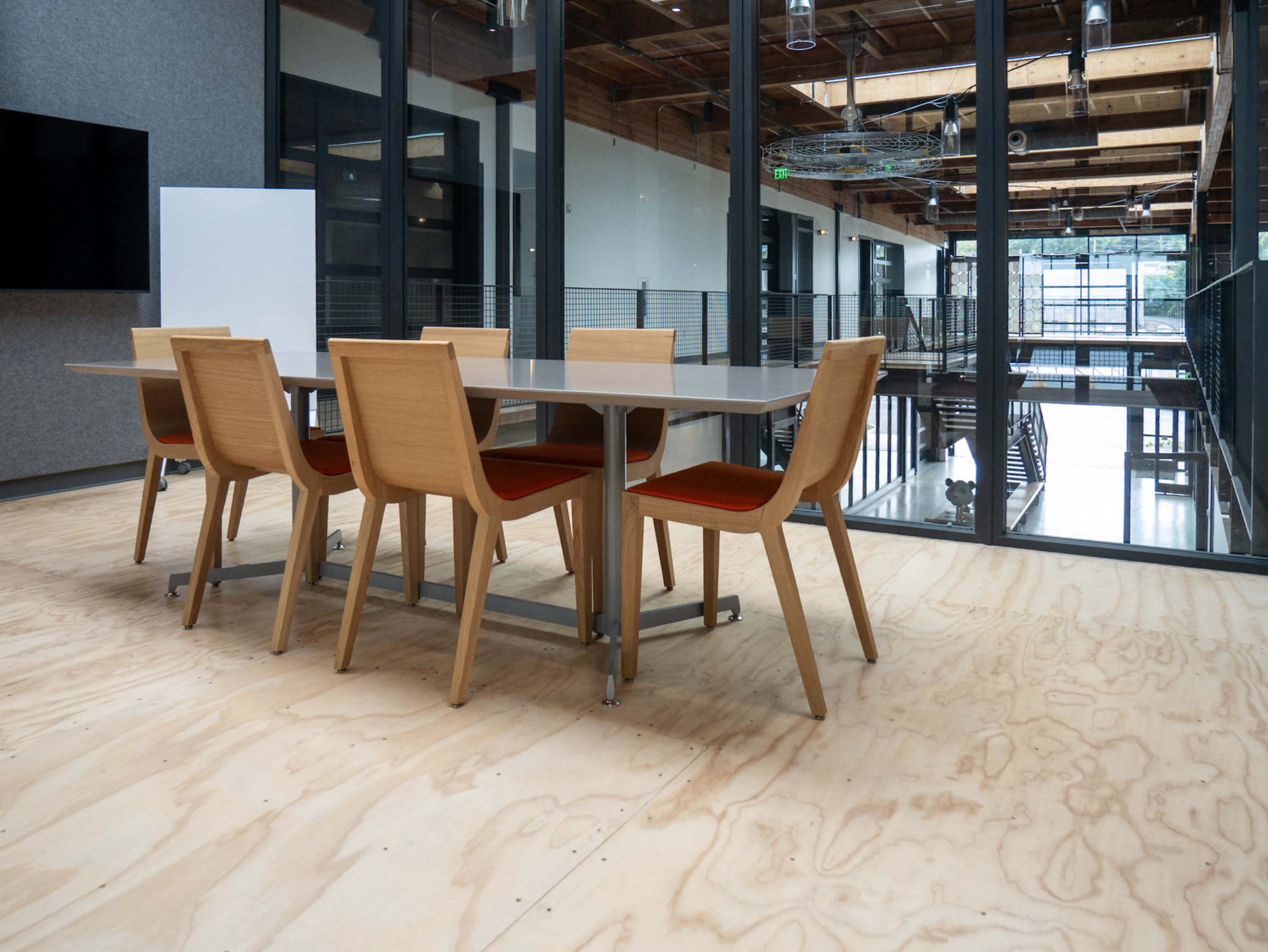 A modern conference room features a large wooden table surrounded by six wooden chairs on a plywood floor.
