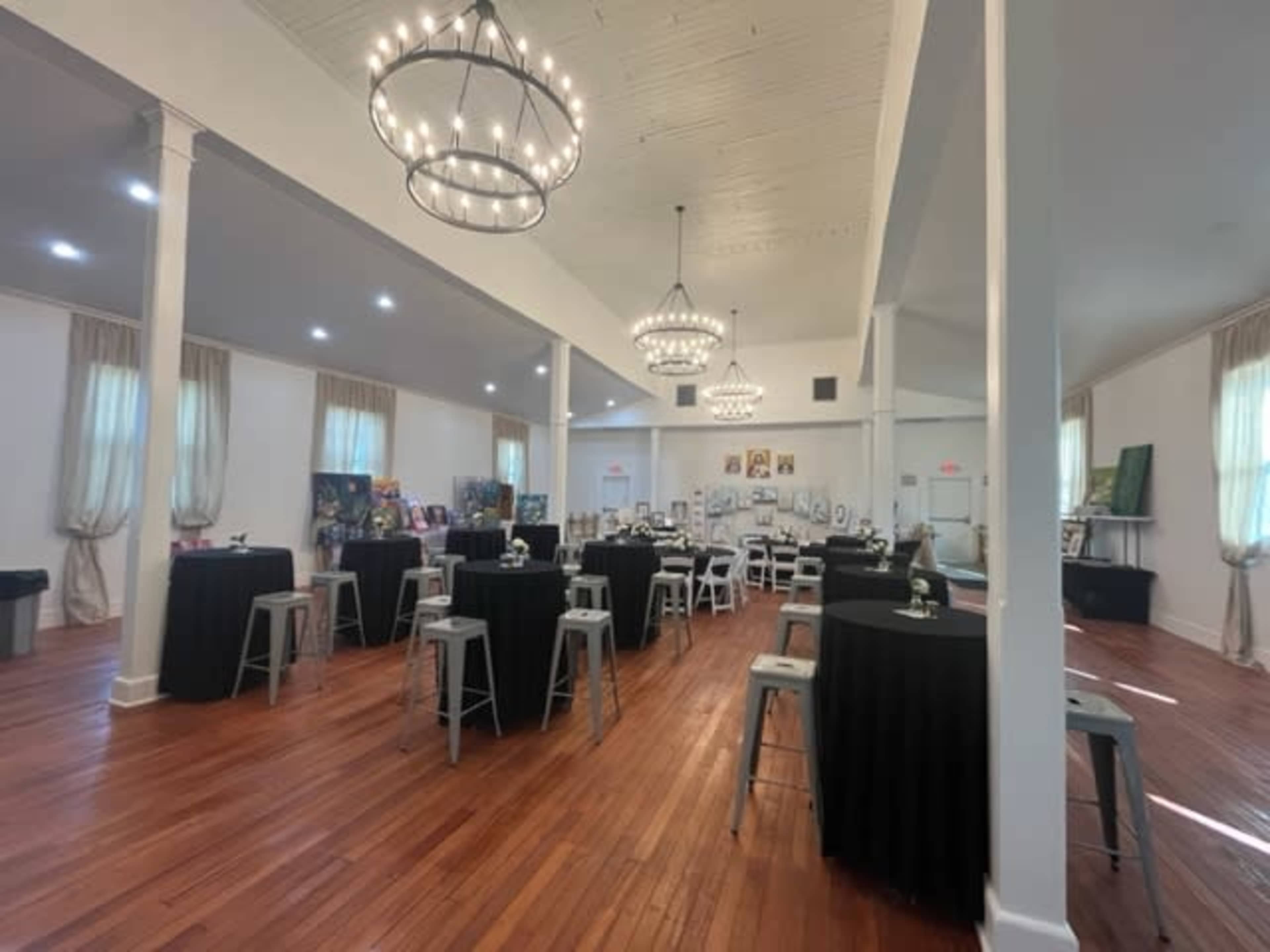The image shows a spacious event hall with wooden flooring, round tables covered in black tablecloths, and chandeliers hanging from the ceiling.