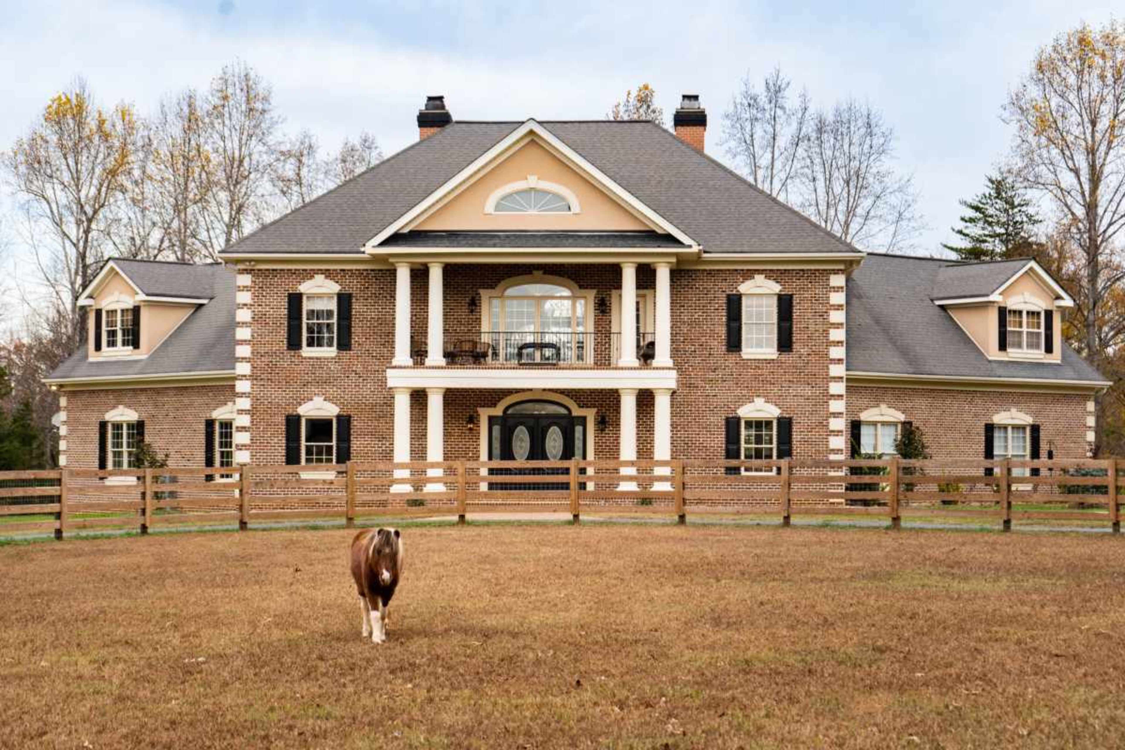 A large brick house with multiple columns and a front porch is situated on a grassy plot, with a cow grazing nearby and trees in the background.