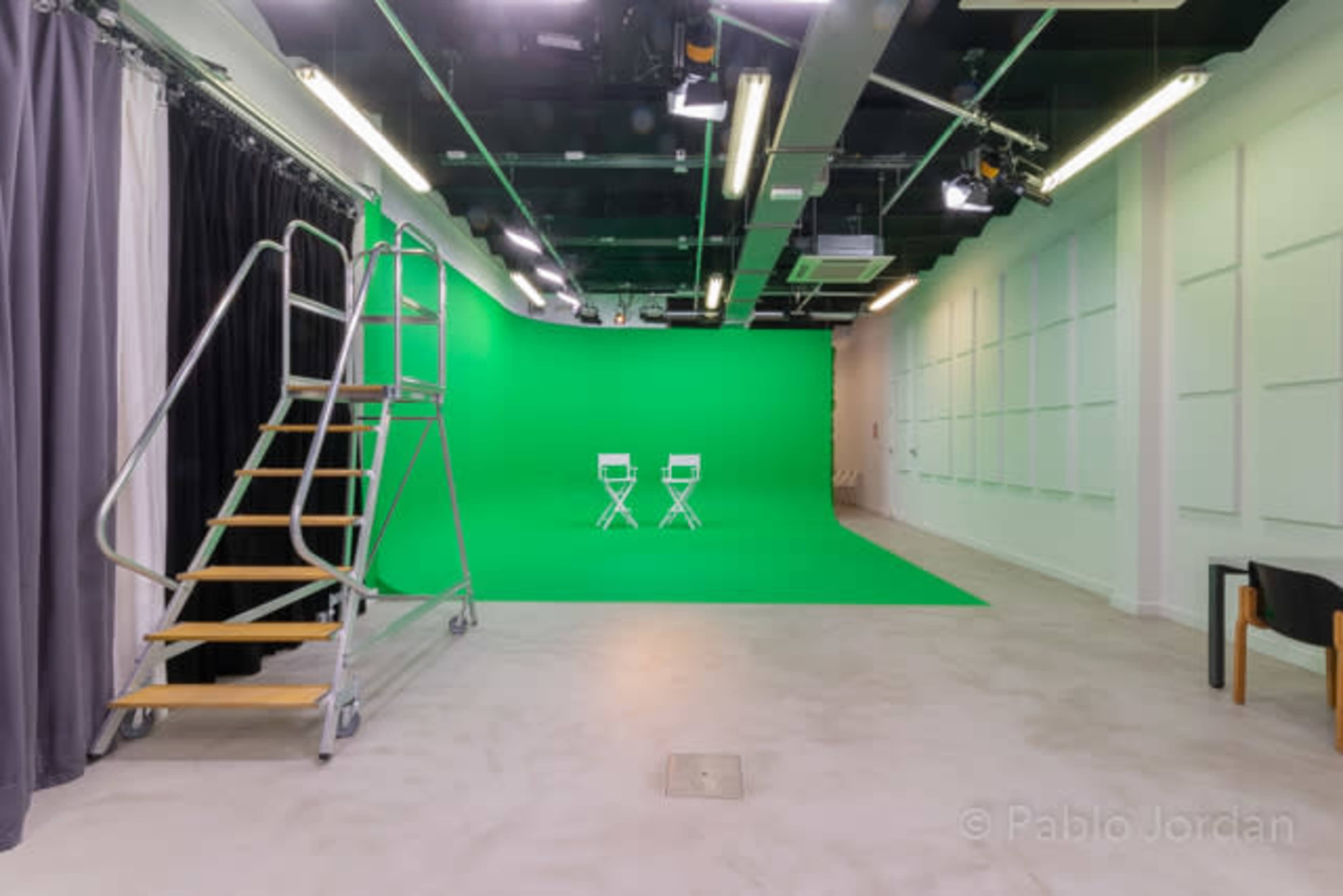 A studio with a green screen in the background, two folding chairs set on the green surface, and a ladder against the wall.