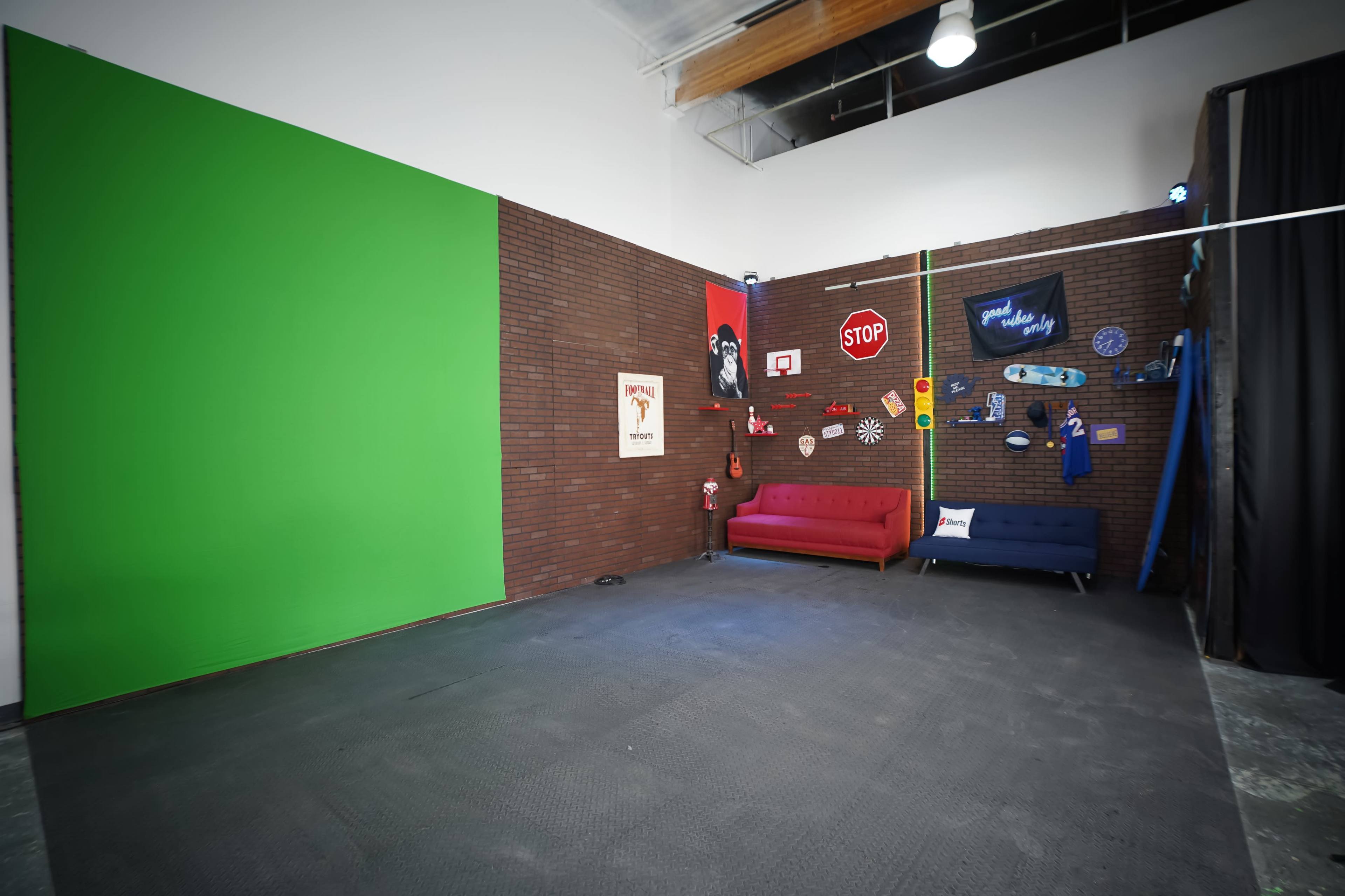 The scene features a studio space with a green screen on one wall, two sofas in red and blue, and various decorative items hanging on a brick wall.