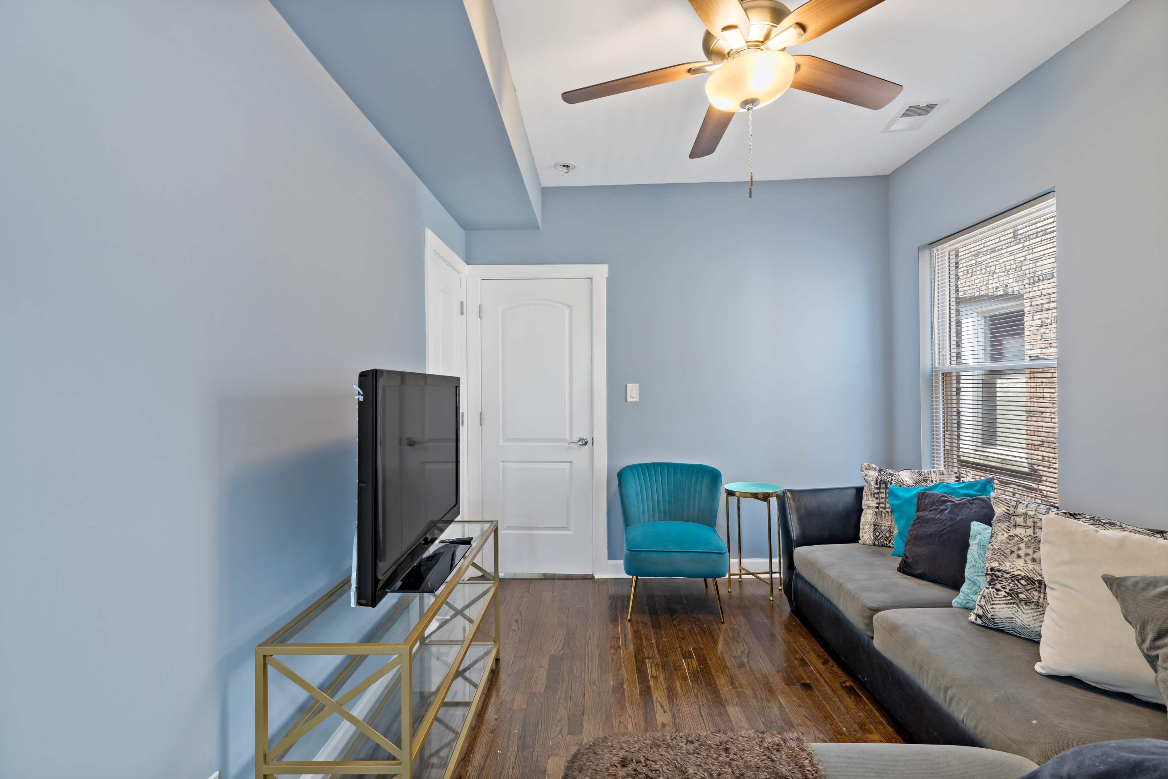 A cozy living room featuring a TV on a gold frame stand, a blue chair, a sectional sofa with decorative pillows, and a ceiling fan.