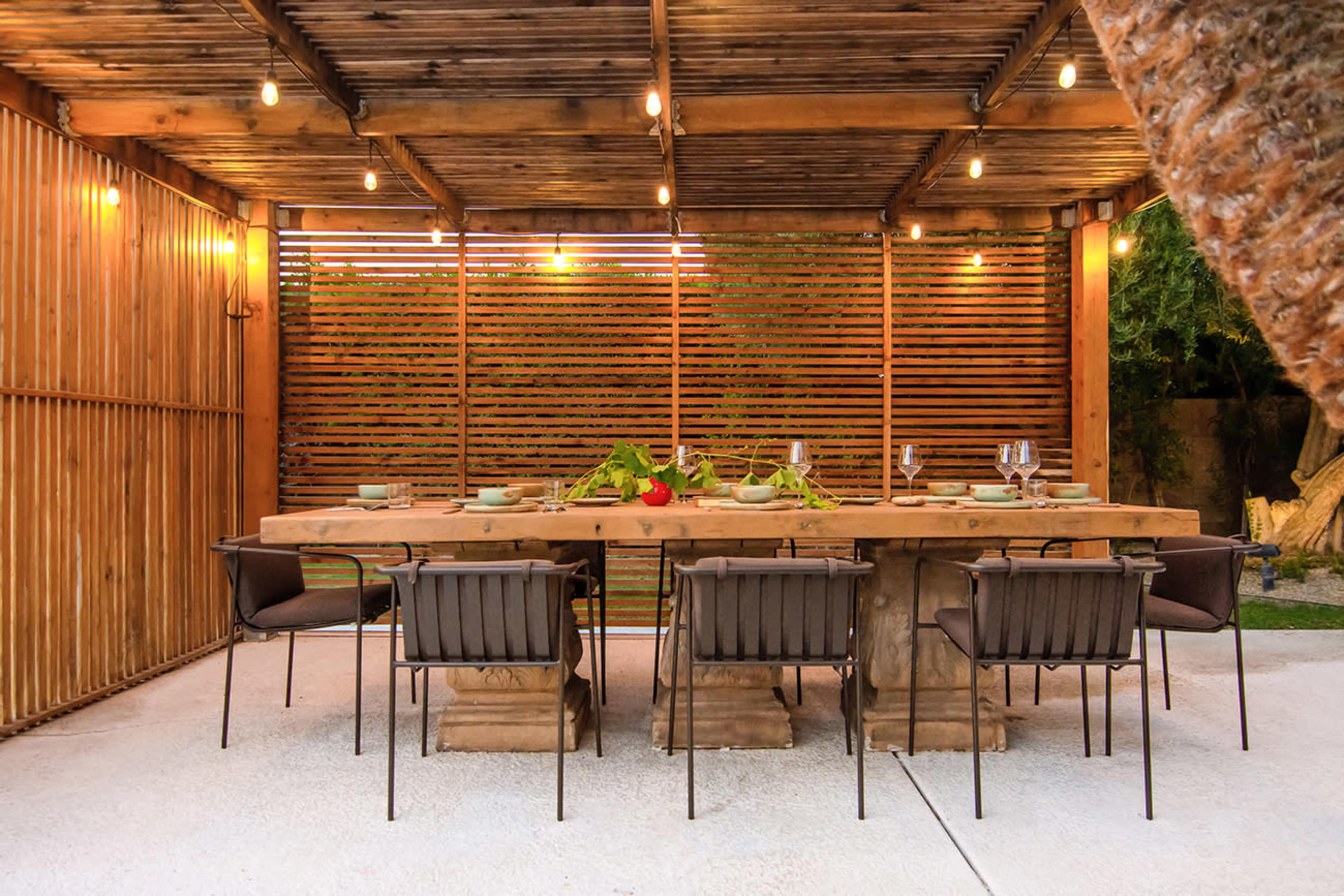 A wooden dining table with several chairs arranged under a pergola adorned with string lights, surrounded by wooden slatted walls.