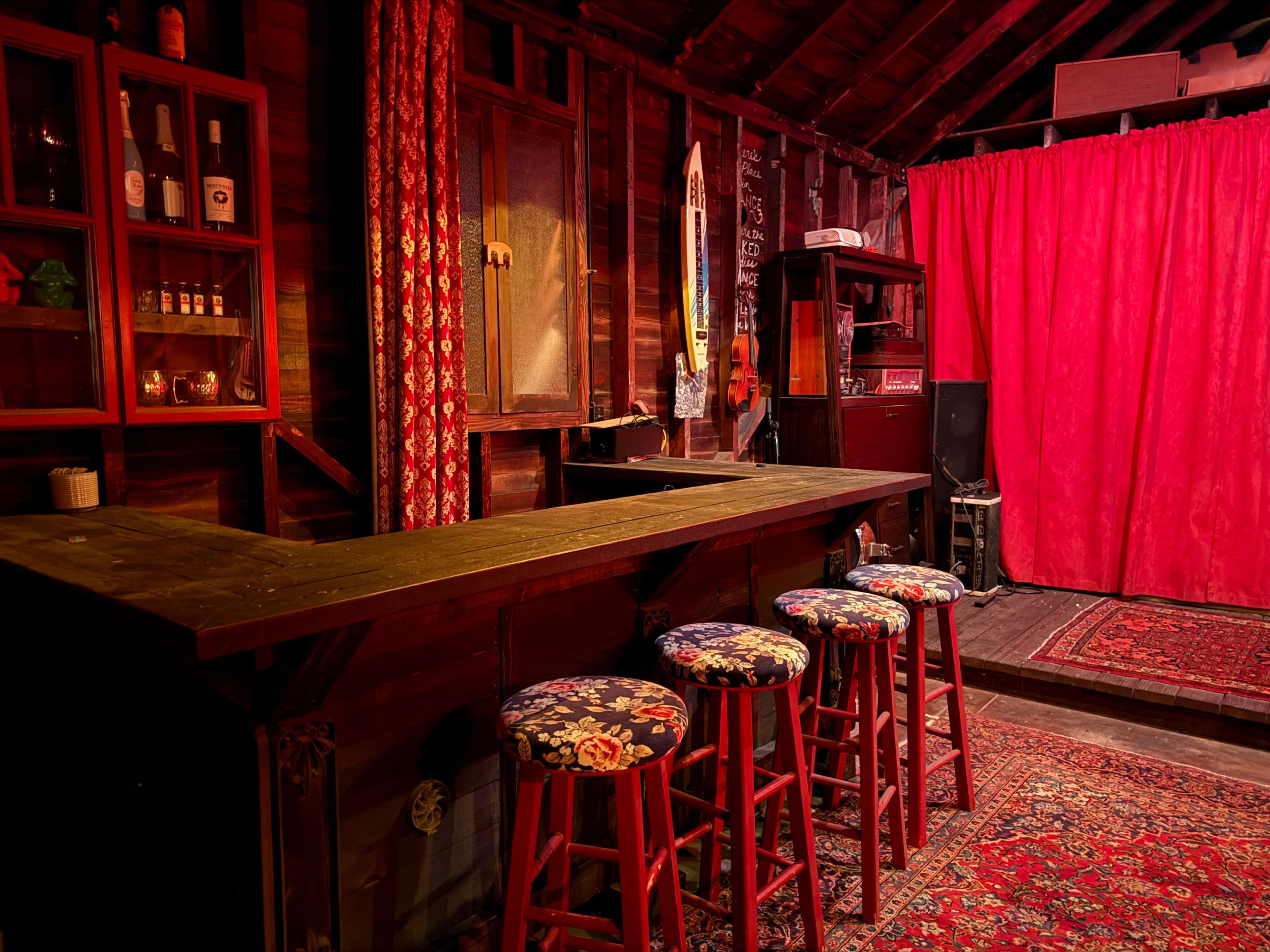 The image shows a rustic bar area with a wooden counter, four upholstered stools, and a backdrop of red curtains inside a dimly lit room.