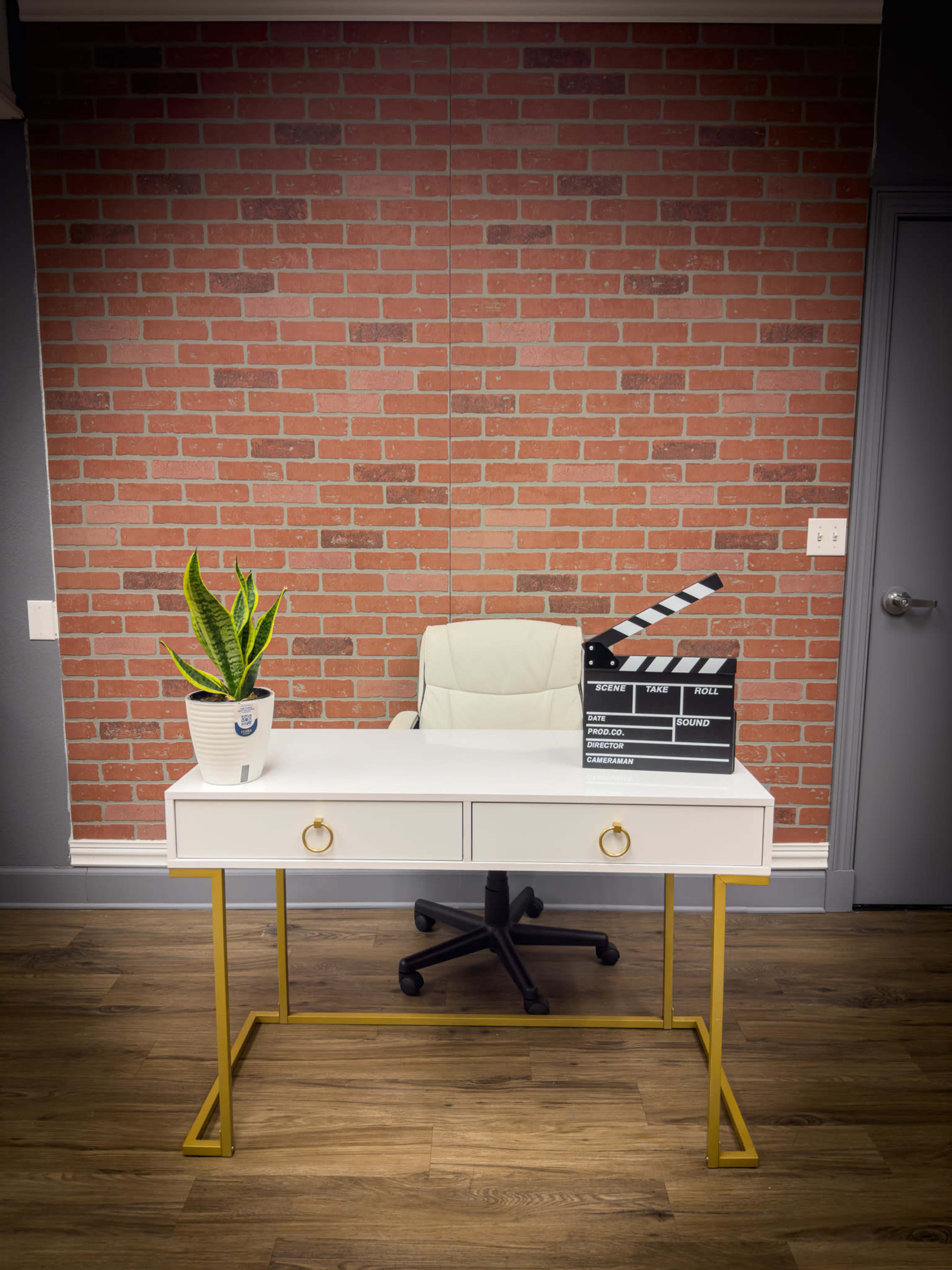 A modern white desk with gold legs is positioned in front of a brick wall, accompanied by a potted plant, a director's clapboard, and an office chair.