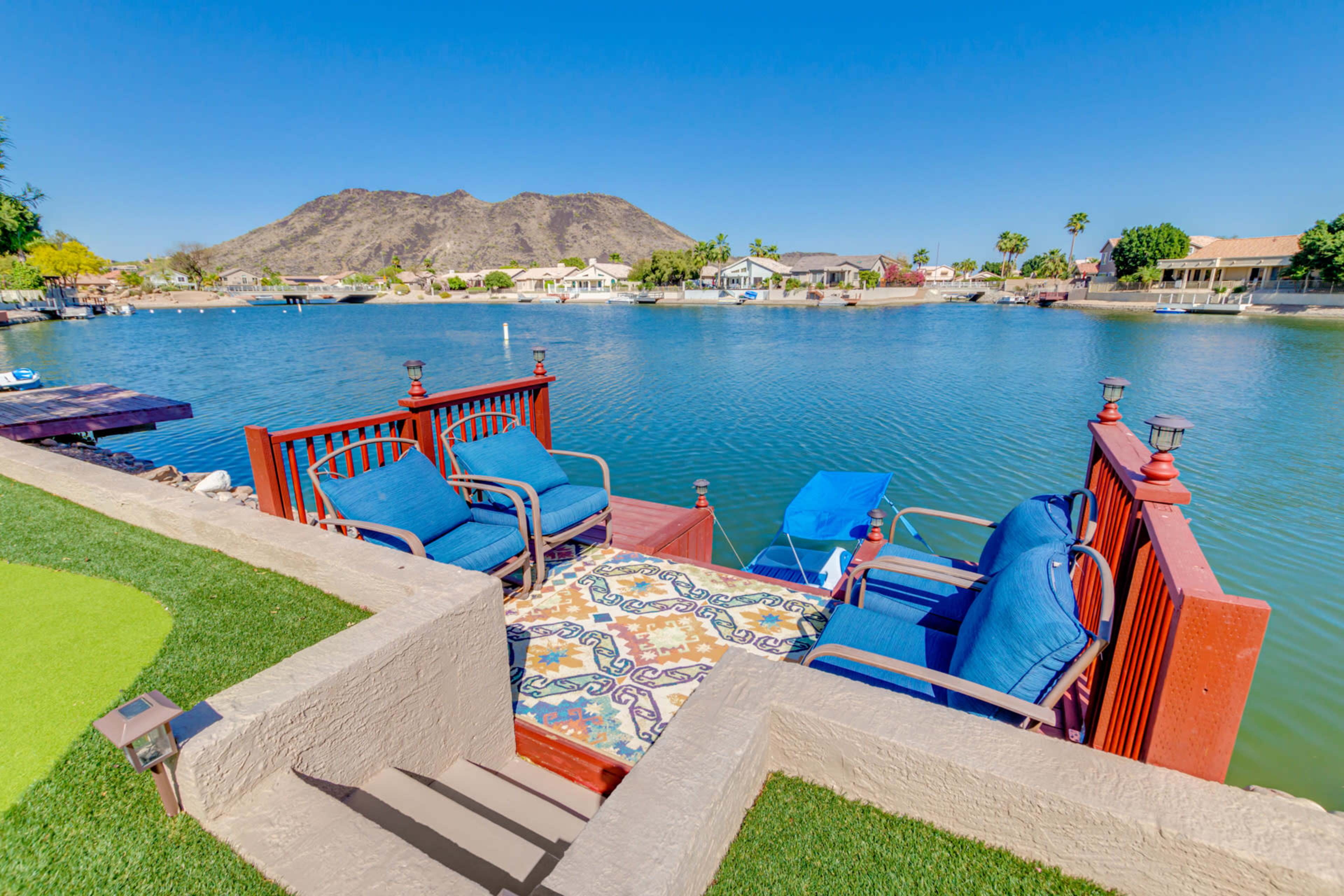 A seating area with blue chairs and a patterned rug overlooks a calm lake bordered by houses and a mountain in the background.