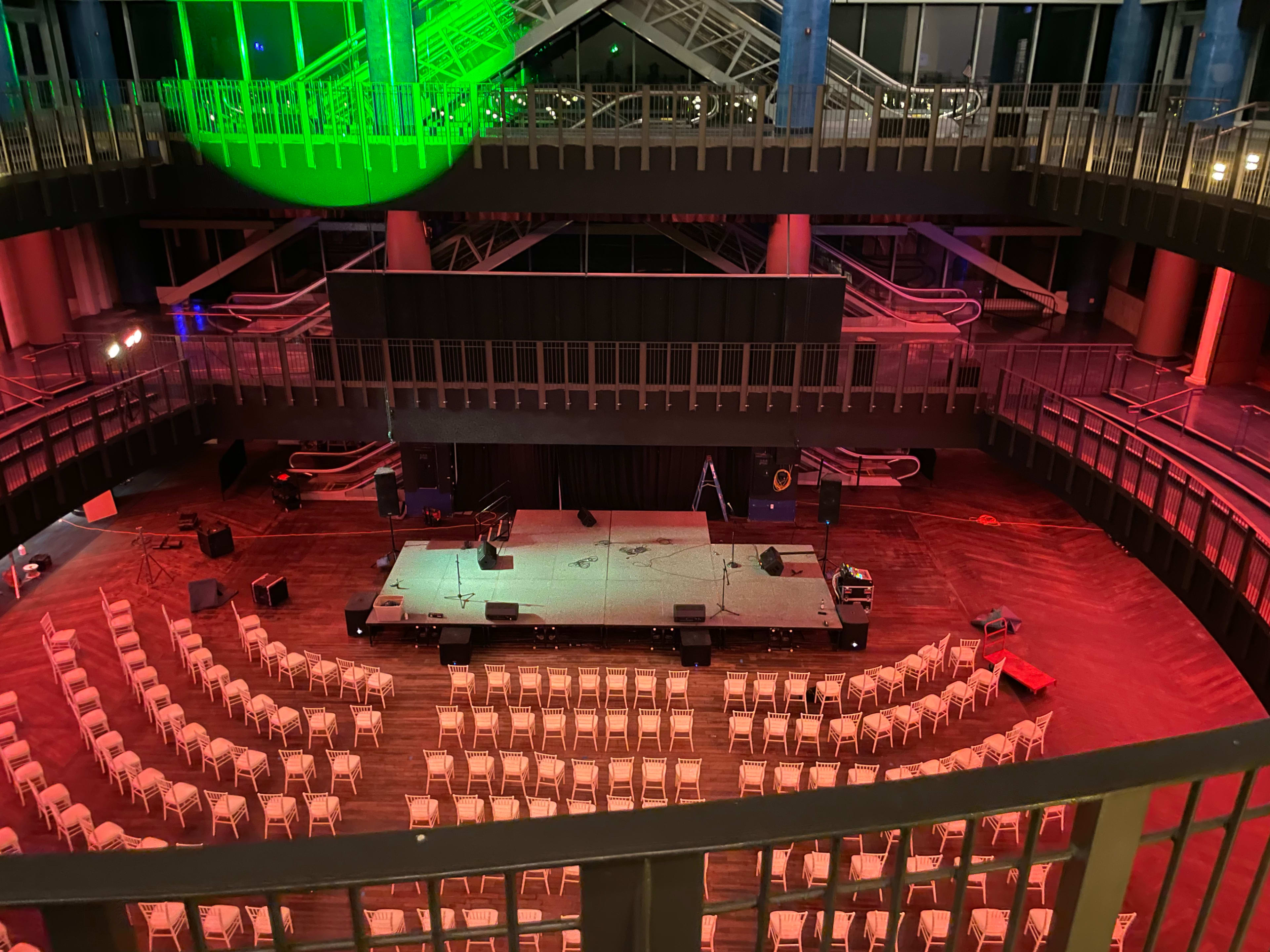 An empty event space with a stage set up at the front, surrounded by rows of white chairs and colorful lighting effects.