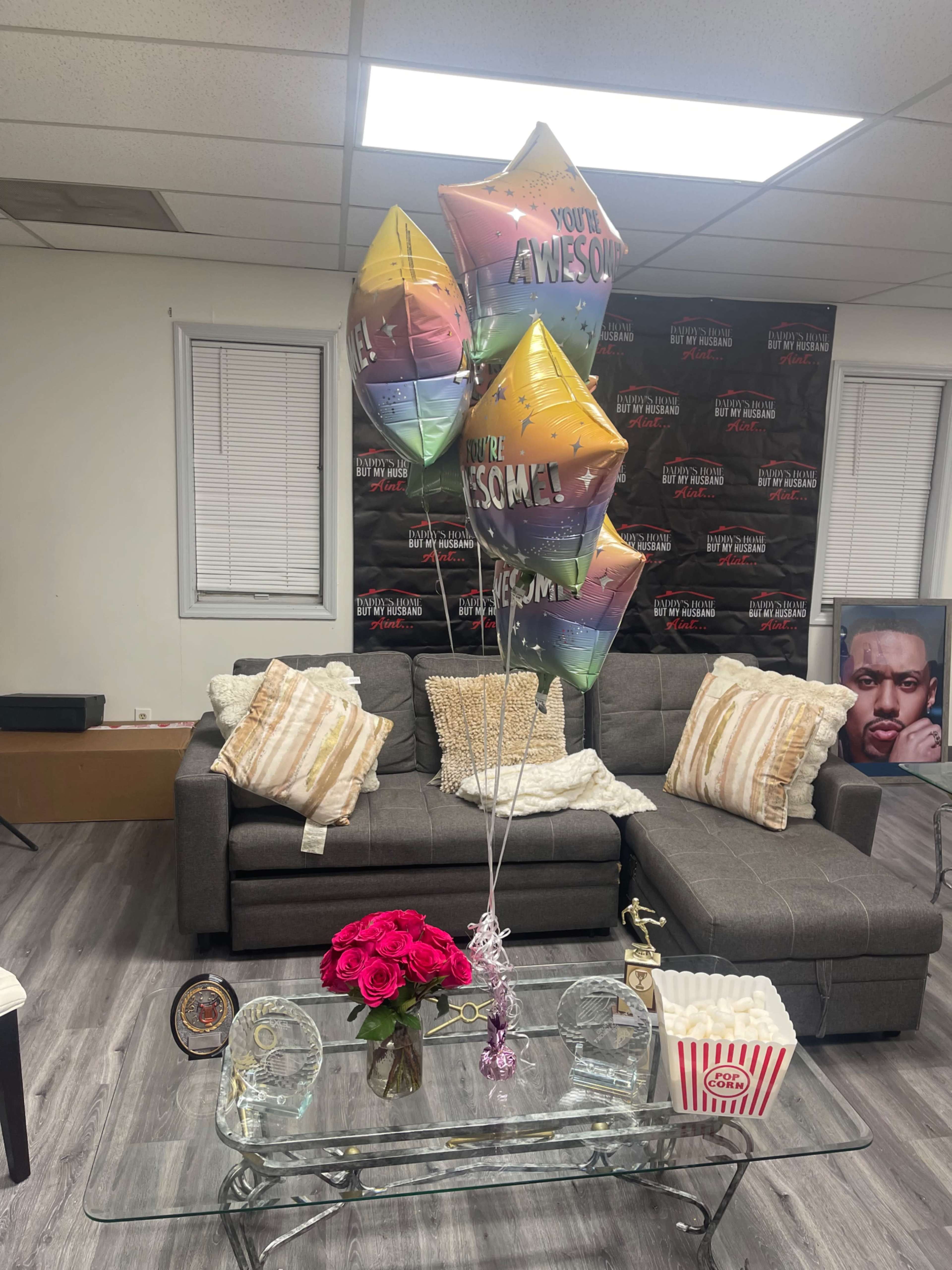 The image shows a cozy living room with a gray sofa, decorative pillows, a glass coffee table adorned with flowers and popcorn, and colorful balloons tied together.