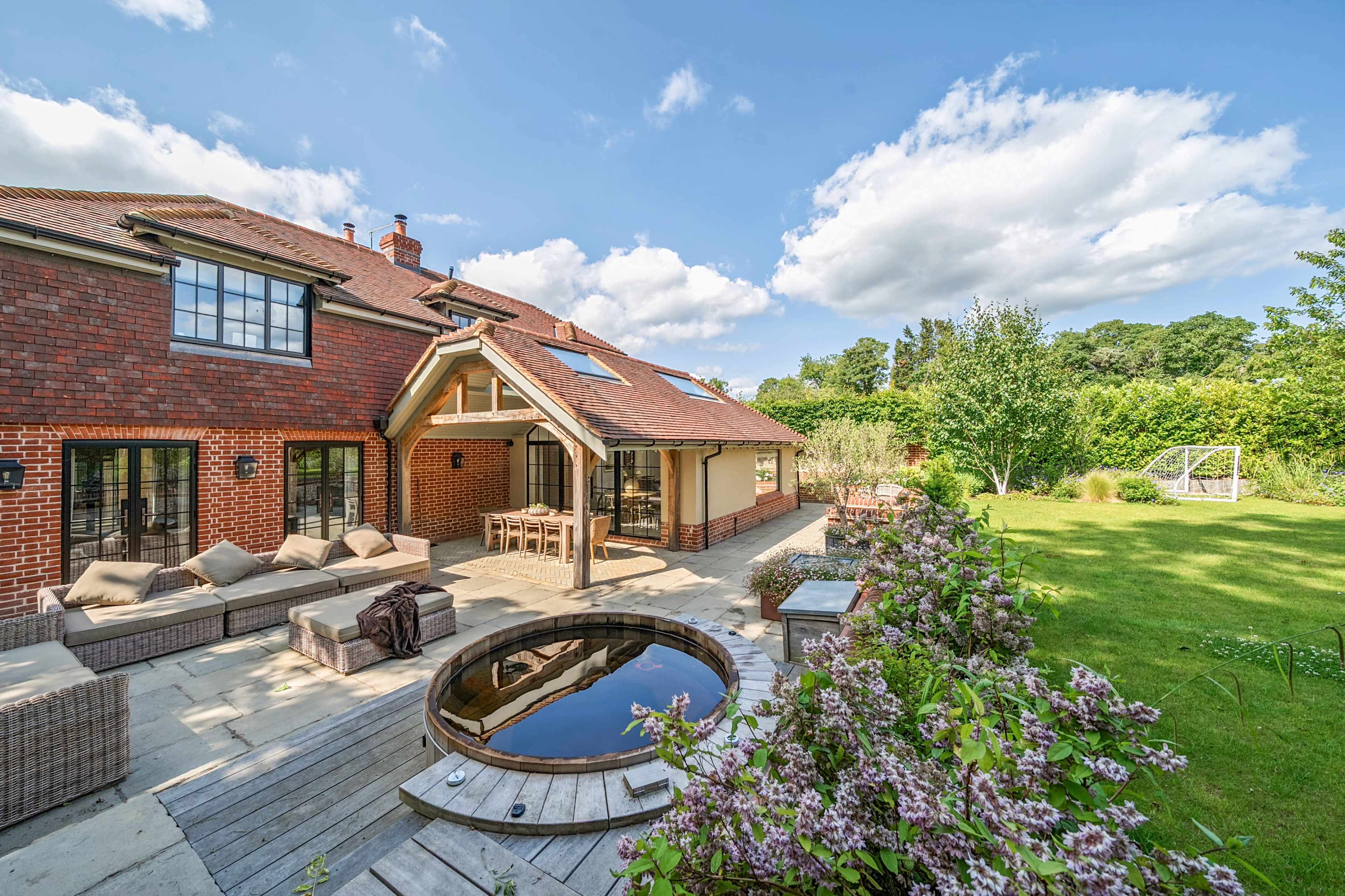 A spacious backyard features a wooden deck with a sunken hot tub, surrounded by blooming flowers and a well-maintained lawn, adjacent to a brick house with large windows.