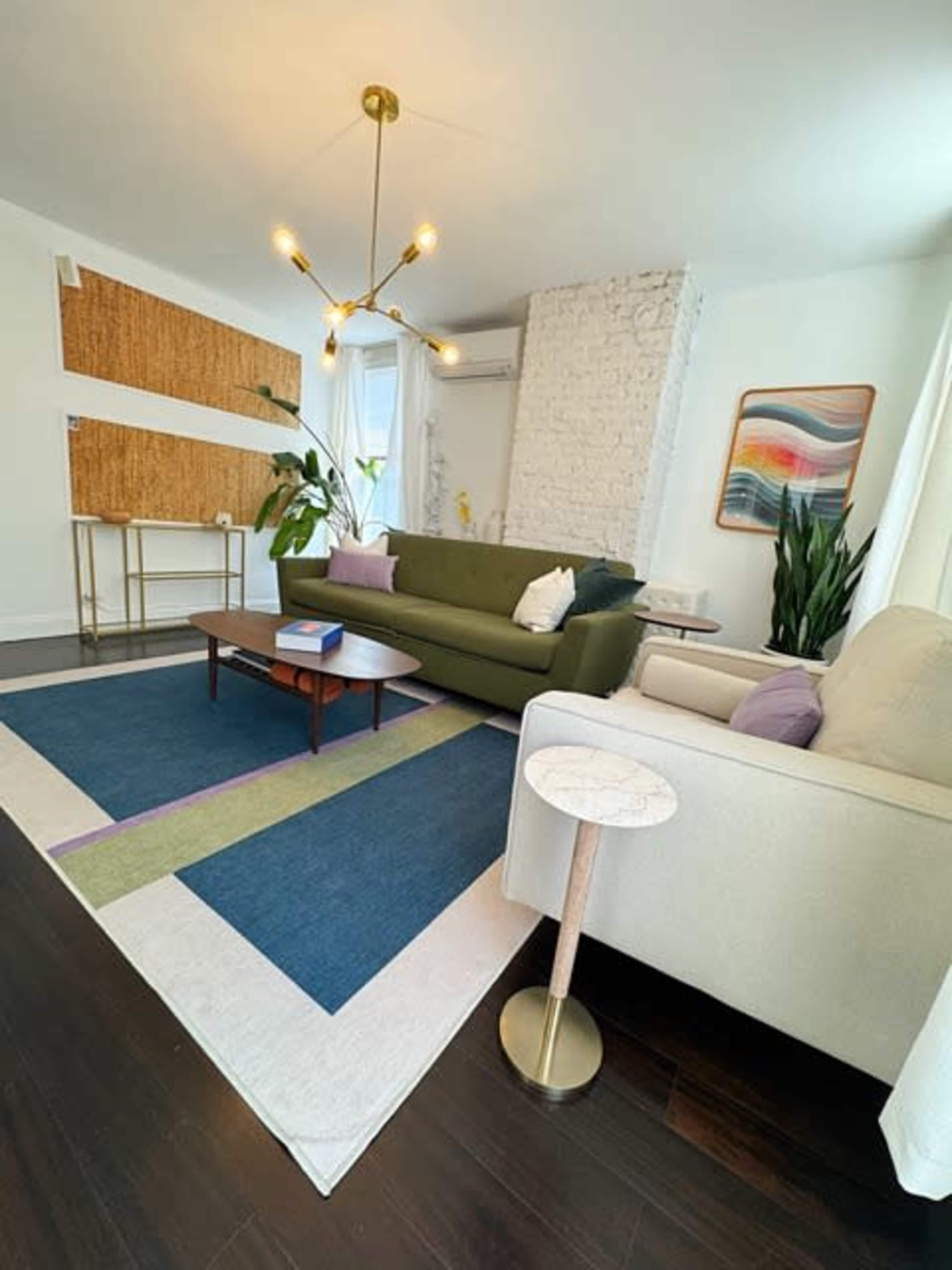 The living room features a green sofa, a white armchair, a large area rug with geometric patterns, and a decorative wall shelf, all arranged around a standing lamp and plants.