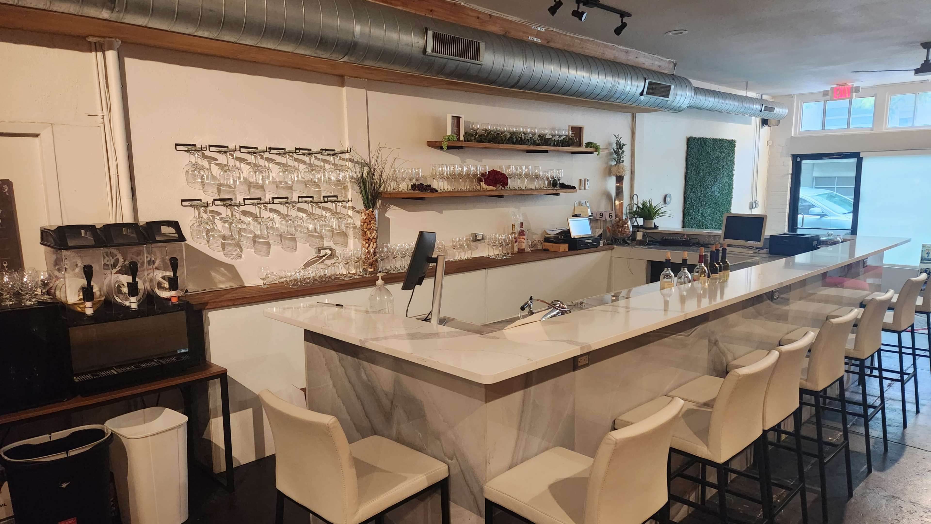 A modern bar area with a marble countertop, tall chairs, and a wall of hanging wine glasses, illuminated by overhead lighting.