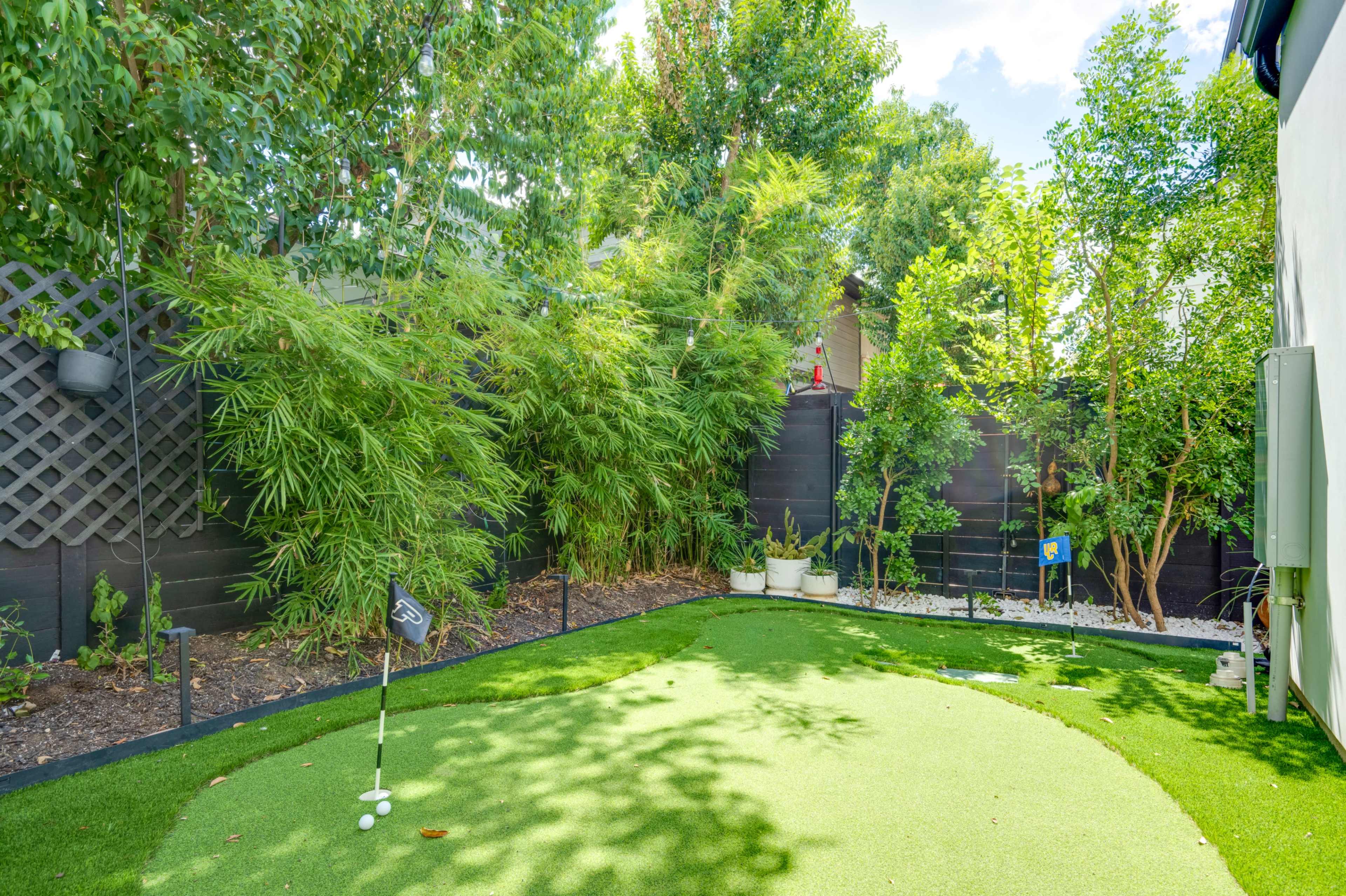 The image shows a small artificial green with a golf hole surrounded by lush greenery and tall plants in a backyard setting.