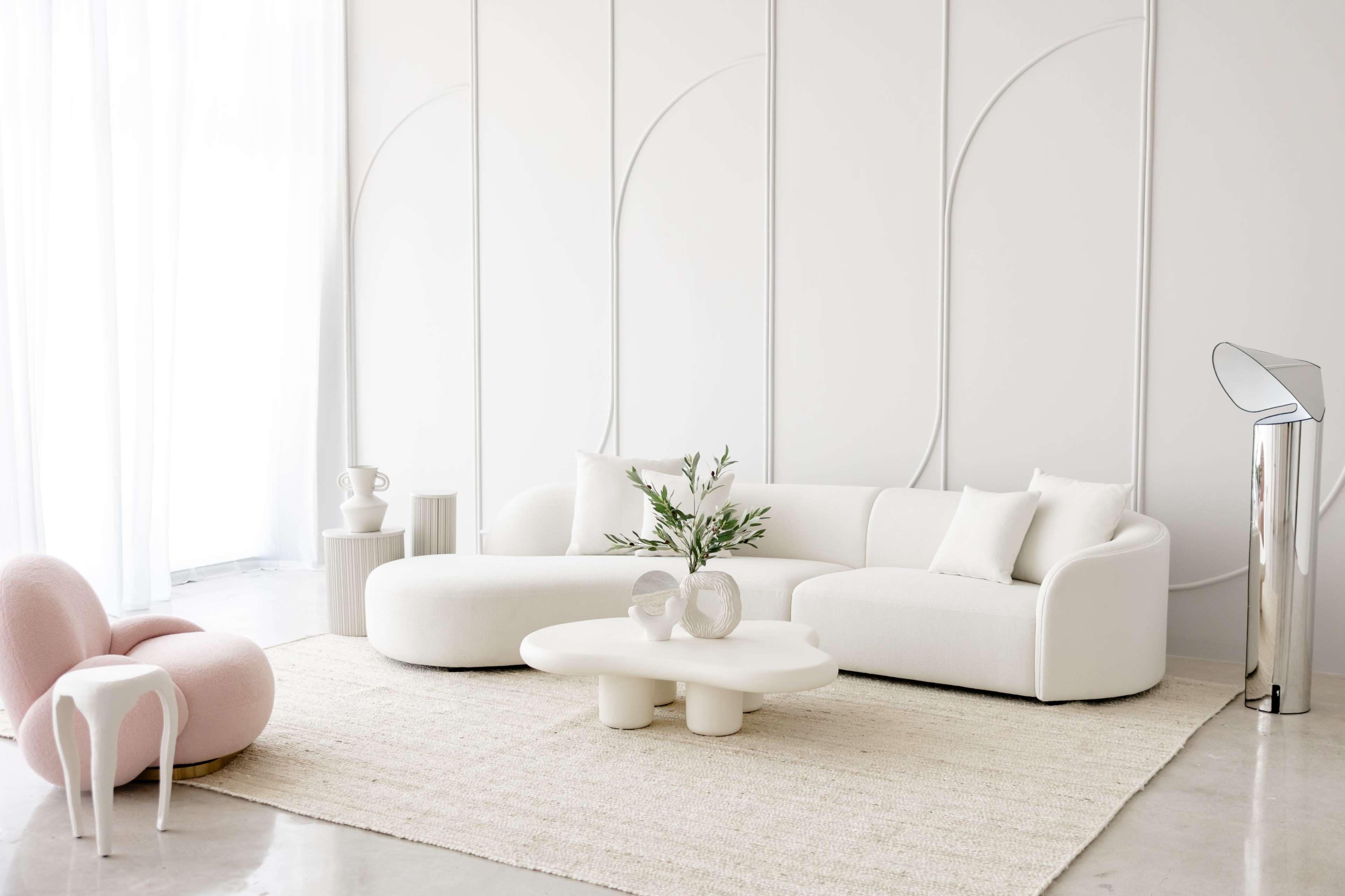The image depicts a minimalist living room featuring a white curved sofa, a circular coffee table, a pink accent chair, and a metal lamp, all set against a white wall with subtle geometric designs.