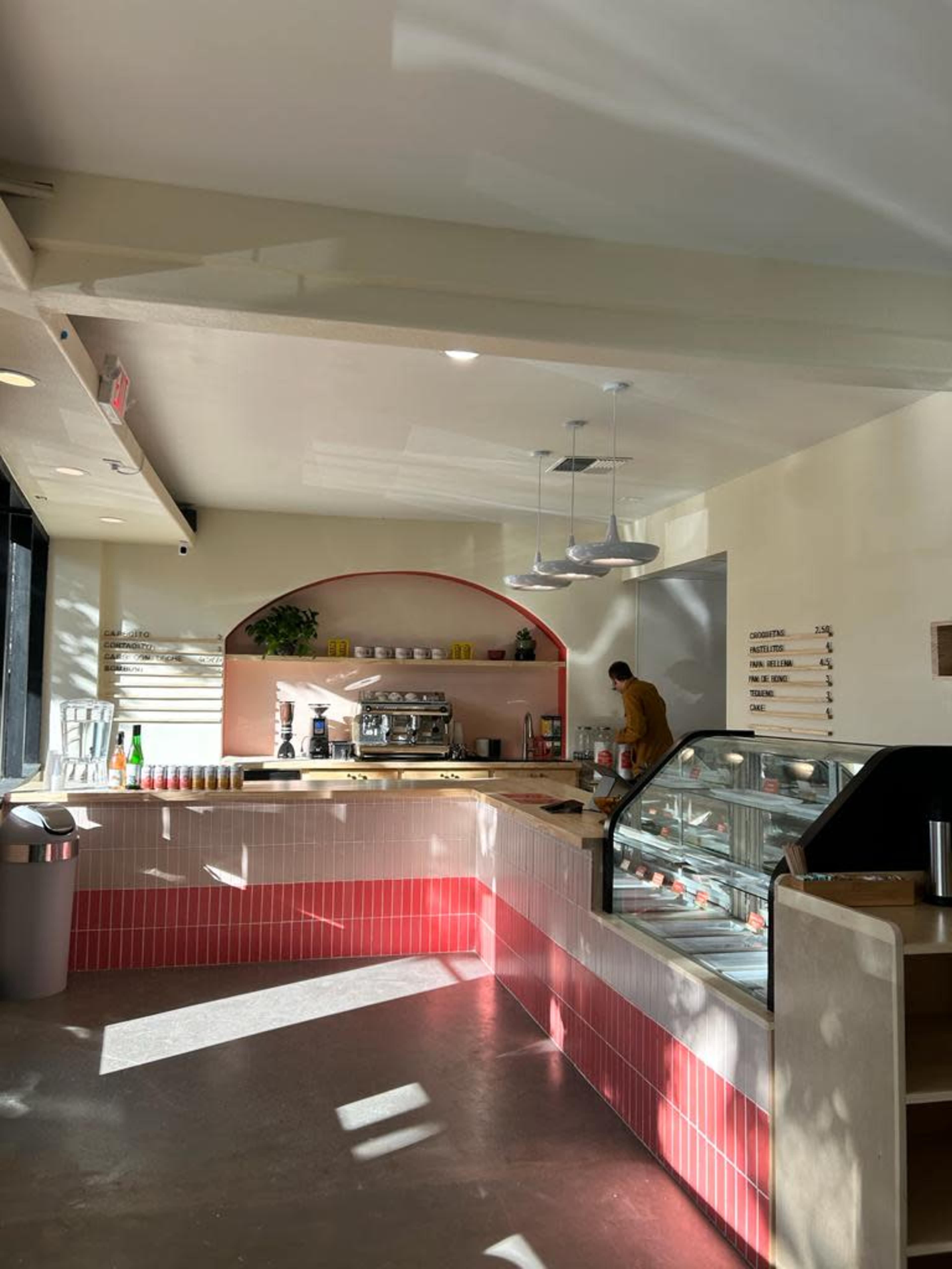 A modern cafe interior features a coffee station, display case, and a worker preparing items under bright overhead lights.