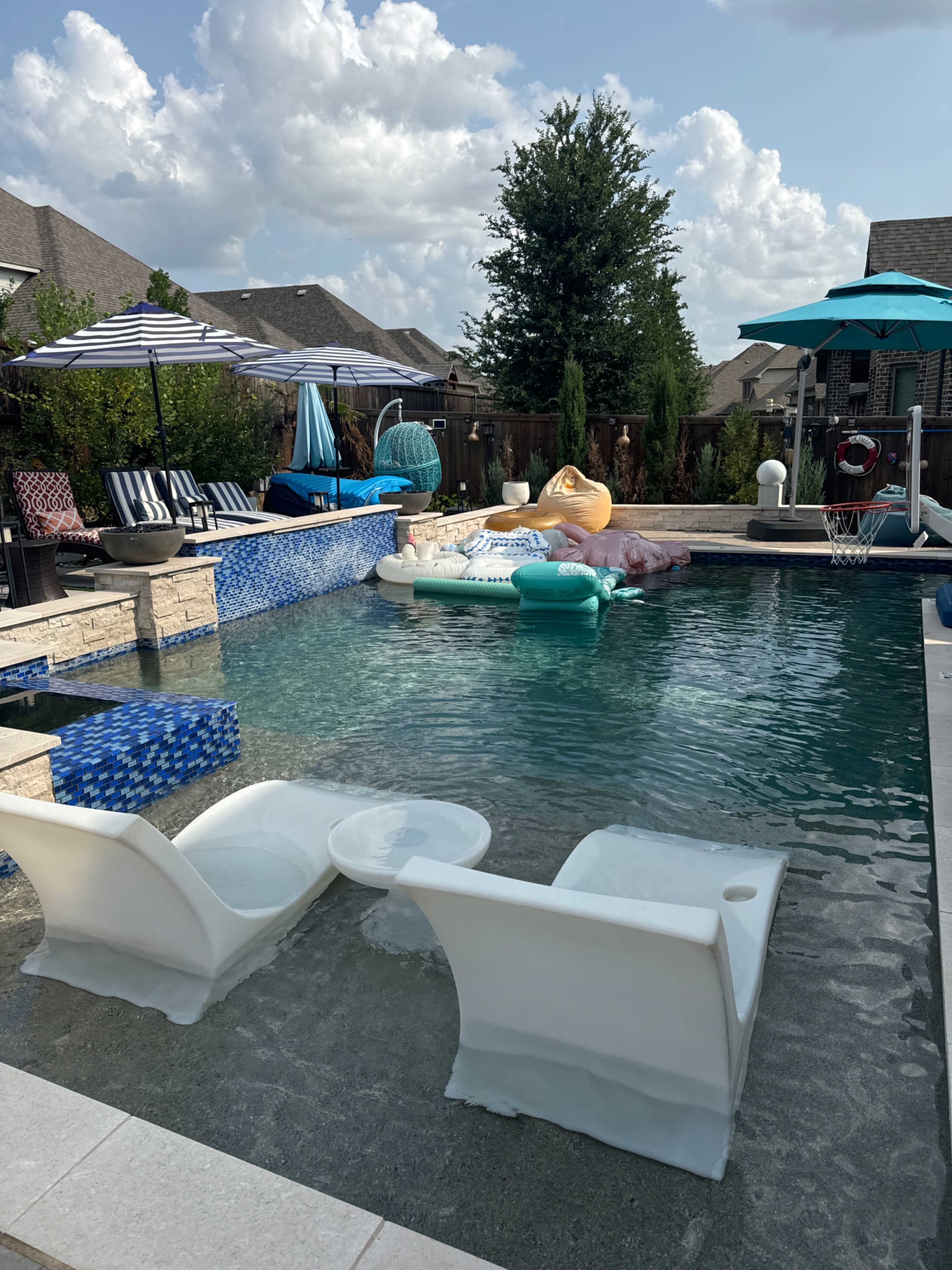 A backyard pool area with lounge chairs, umbrellas, and various inflatable pool floats.