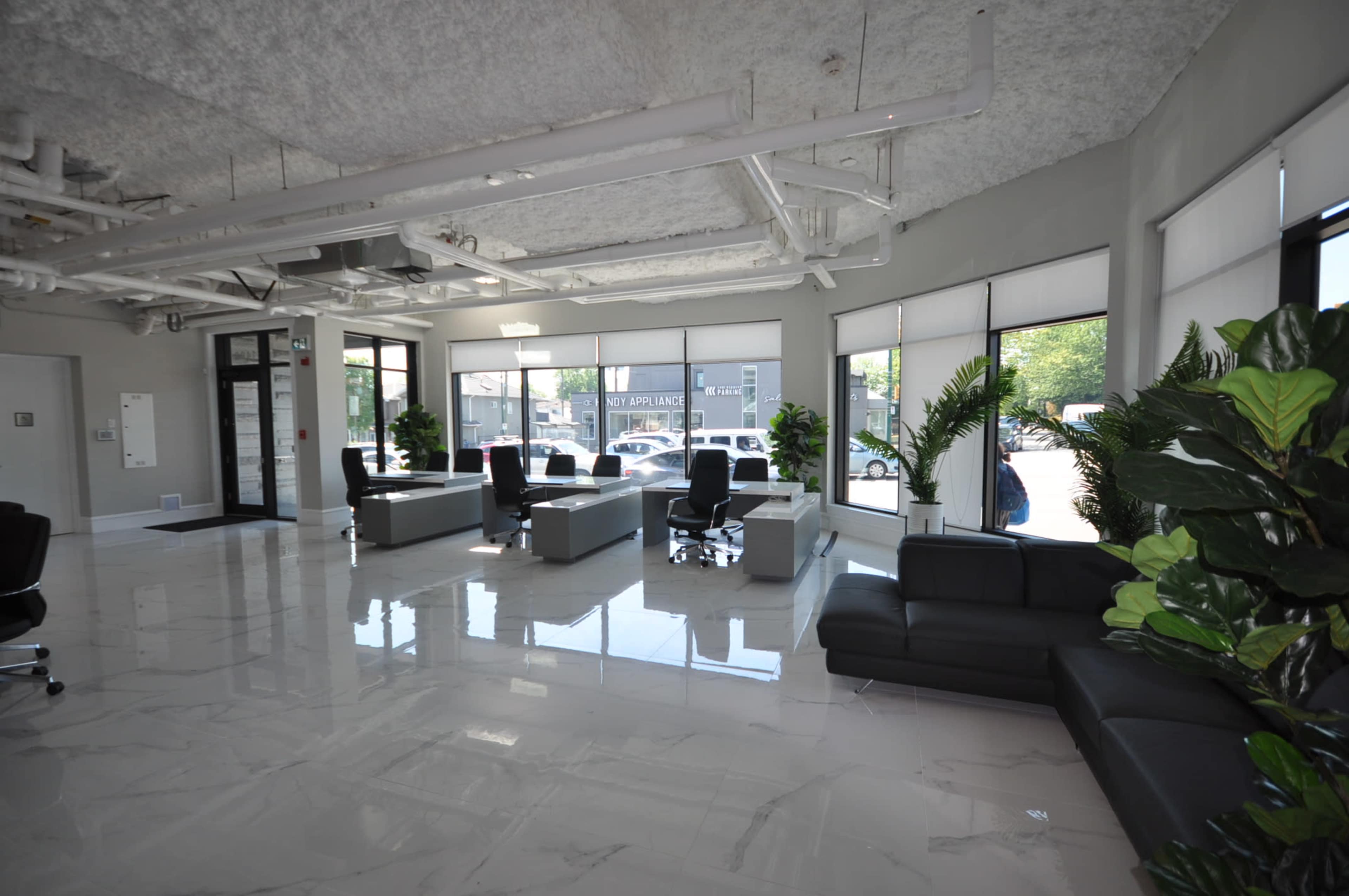 A modern lobby area with gray walls, polished white tile flooring, and multiple seating arrangements, including sofas and office chairs, surrounded by large windows and potted plants.