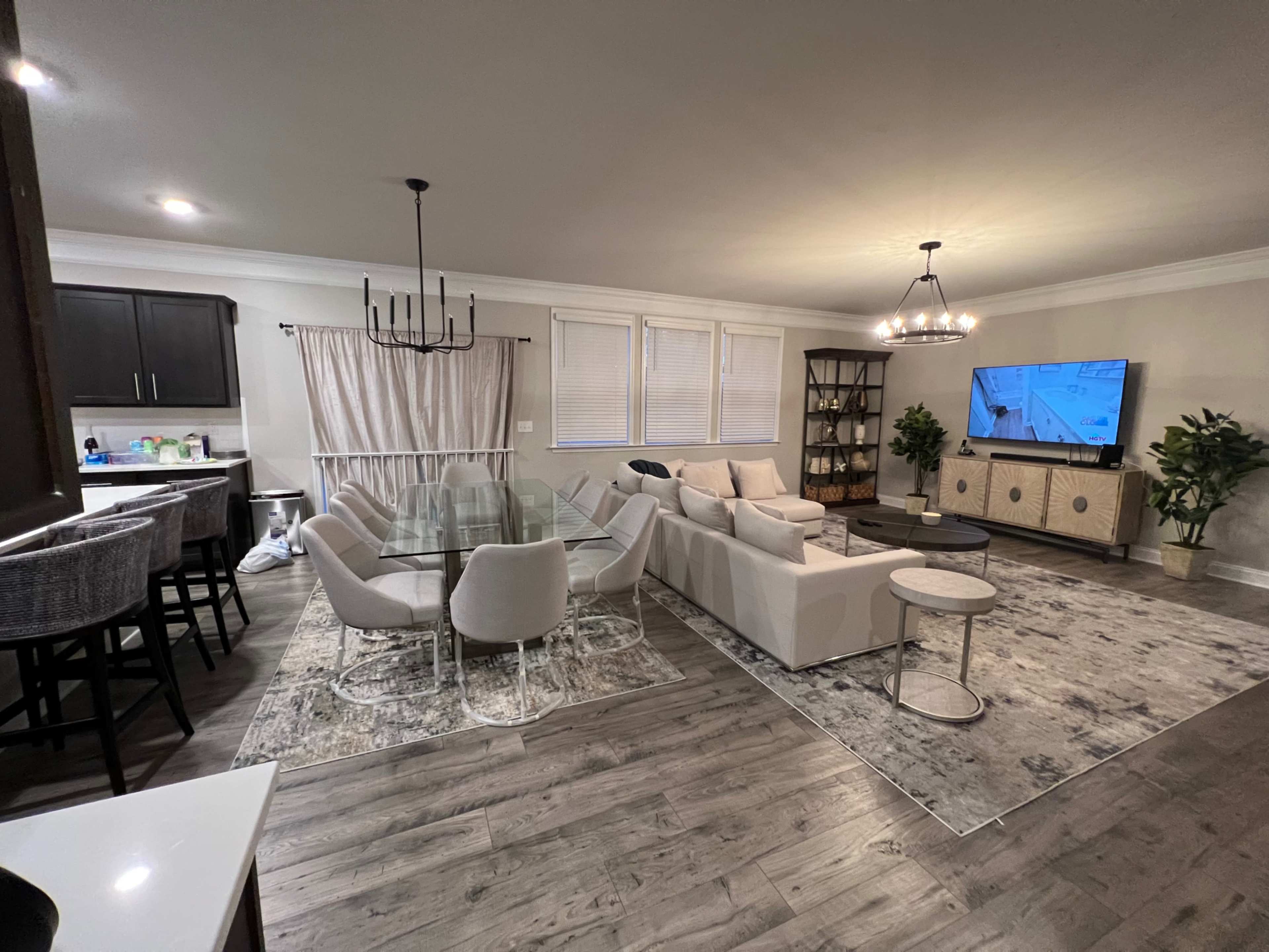 The living room features a glass dining table surrounded by upholstered chairs, a sectional sofa, a TV on a stand, and a decorative bookshelf against the wall.