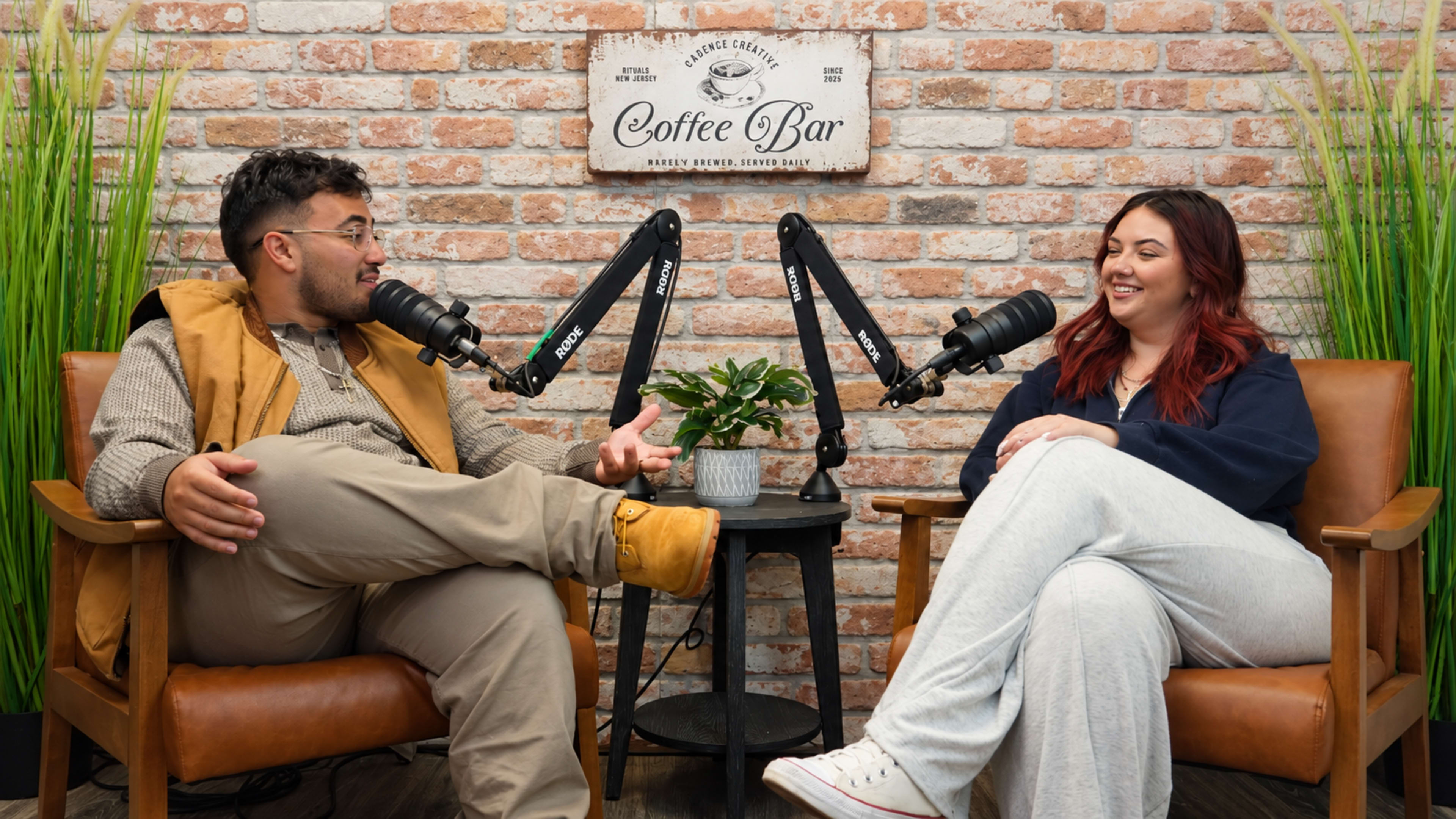 A man and a woman sit in a podcast studio, engaged in conversation while using microphones, surrounded by a brick wall and decorative plants.