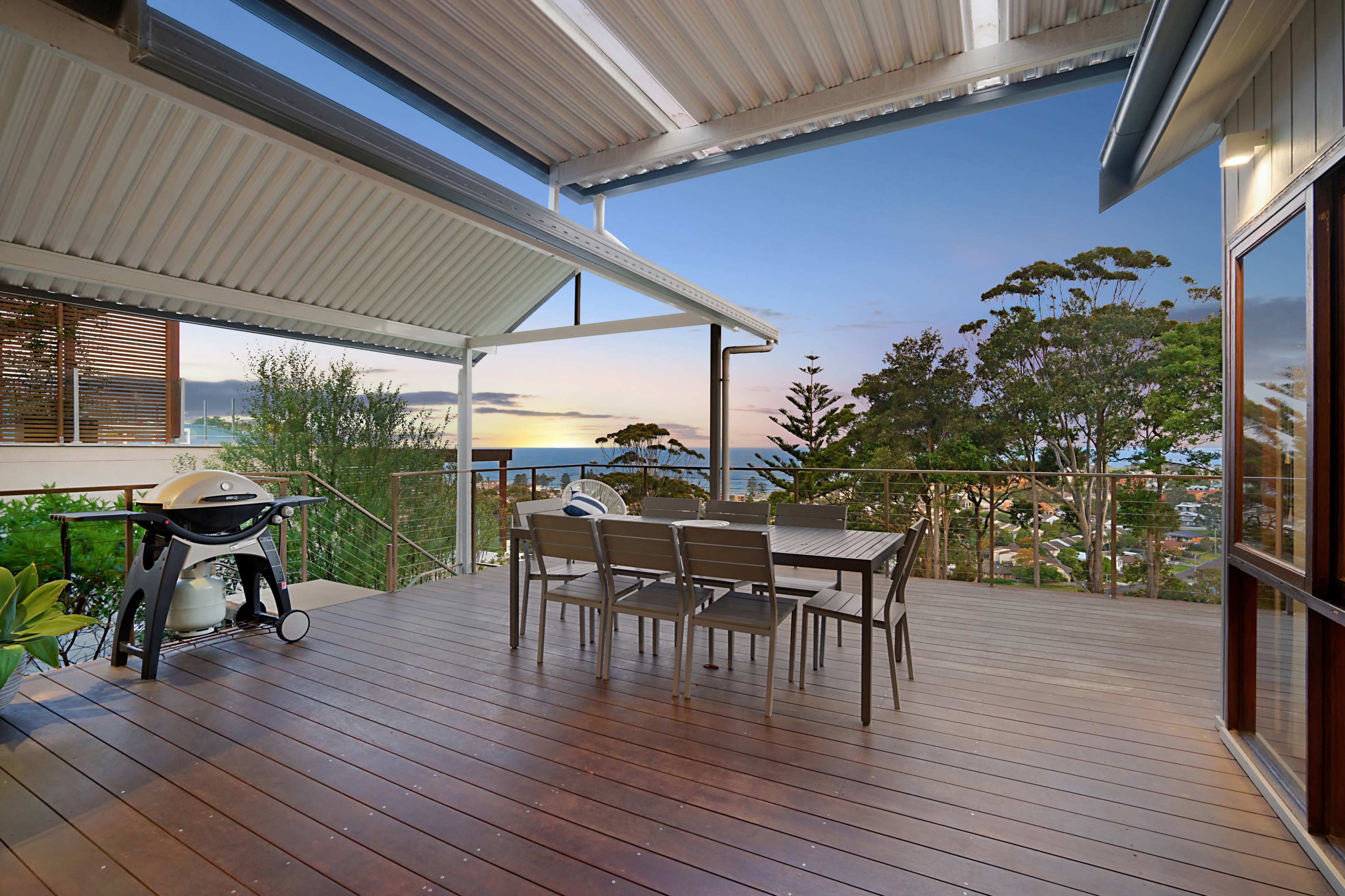 A wooden deck with a dining table and chairs, overlooking a scenic view of trees and a sunset by the ocean.
