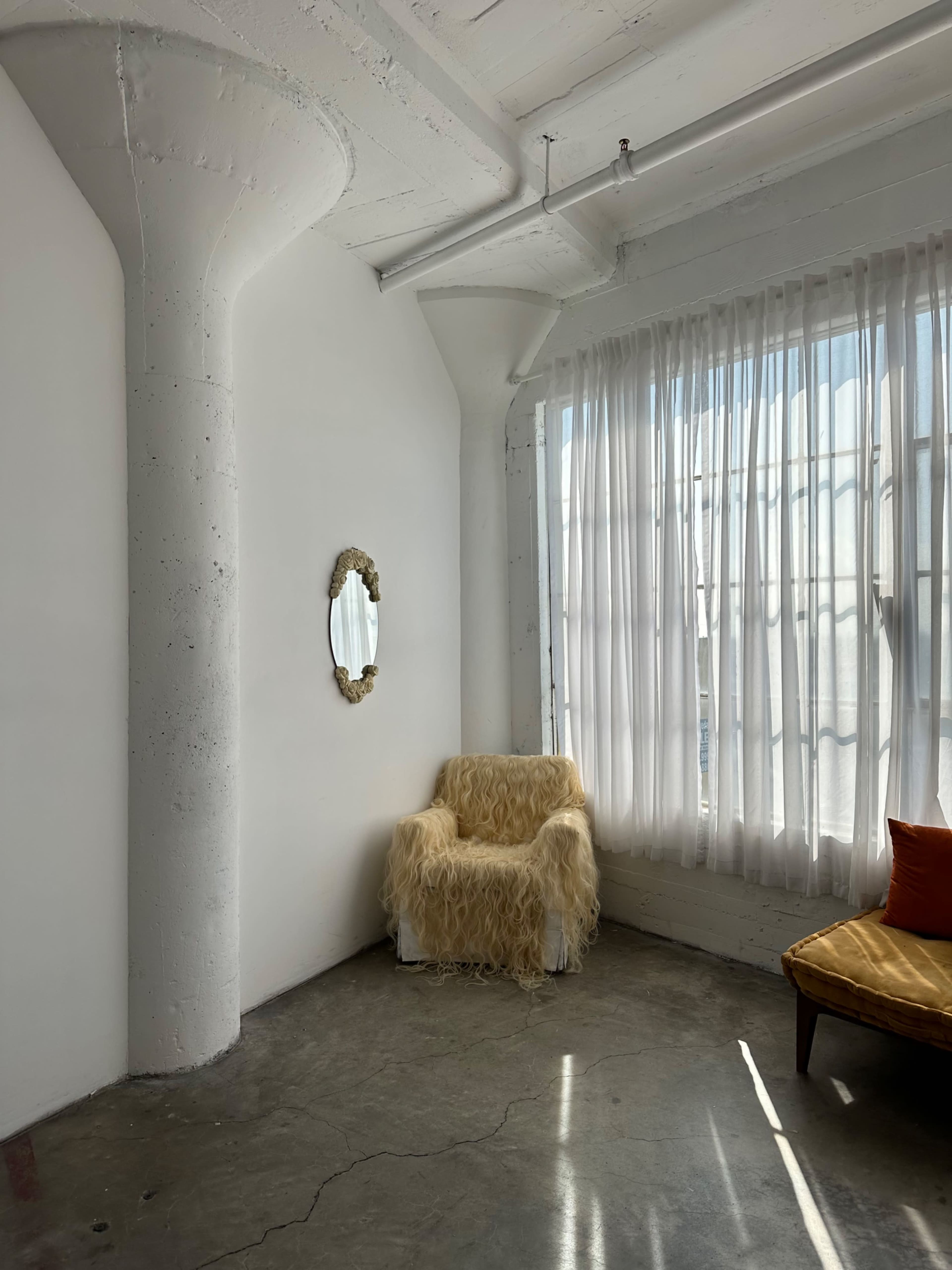 A white-walled room features a plush, shaggy armchair beside a large window with sheer curtains, reflecting a minimalistic aesthetic.