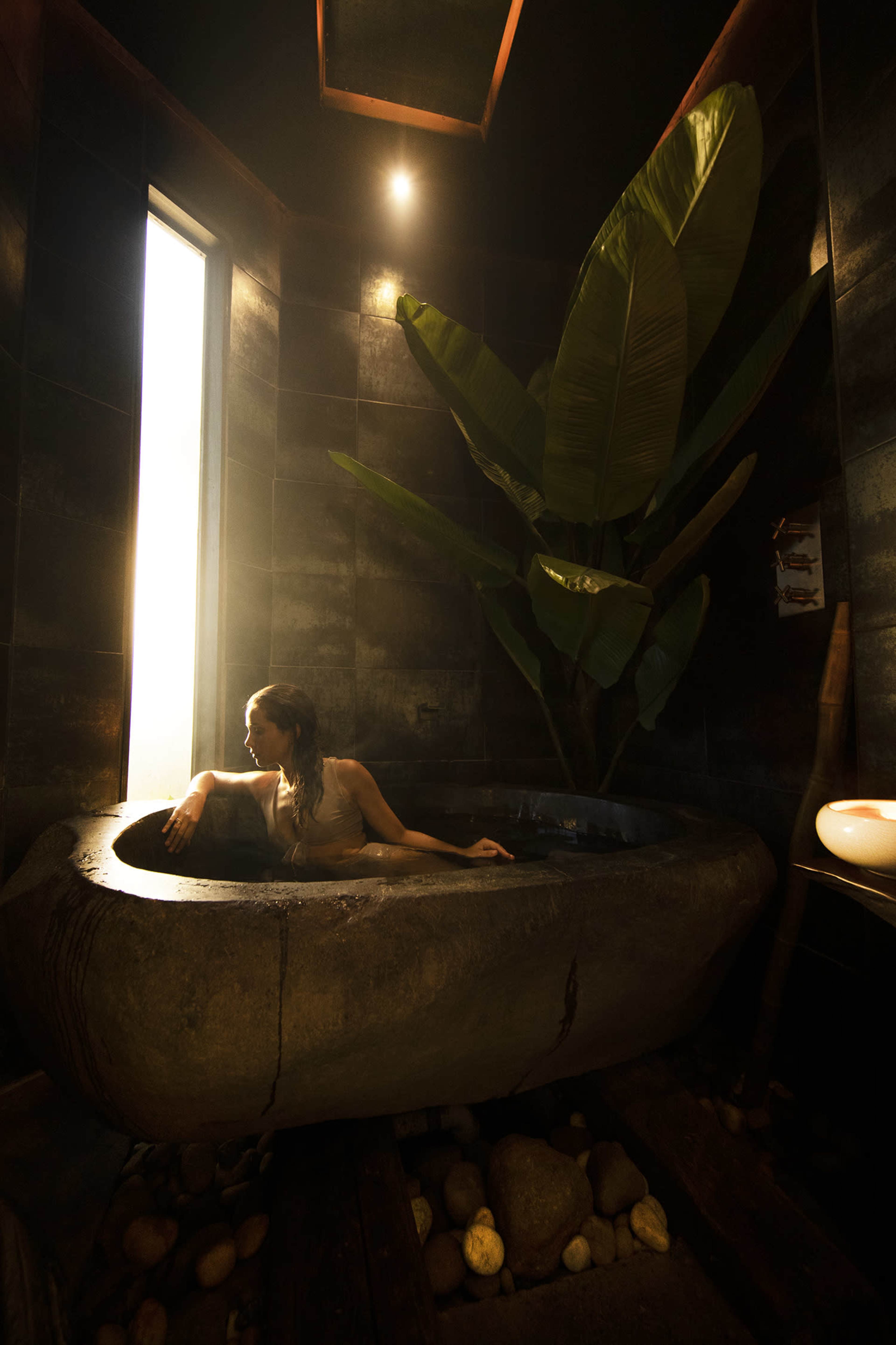 A woman relaxes in a stone bathtub surrounded by dark walls and a large plant near a window.