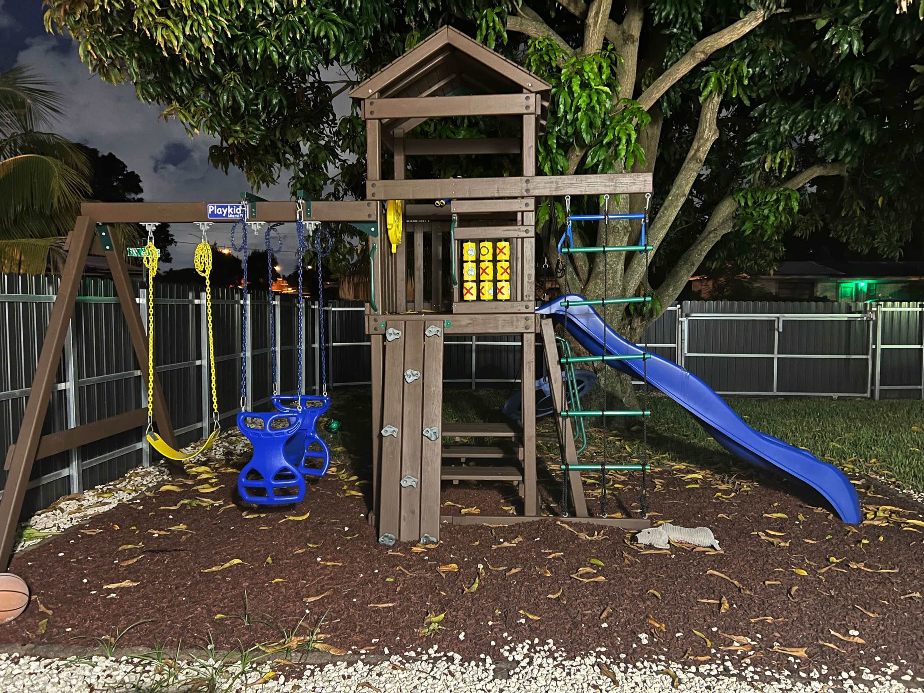 The image shows a wooden playground set with a slide, swings, and a climbing structure, situated on grass and surrounded by a fence.