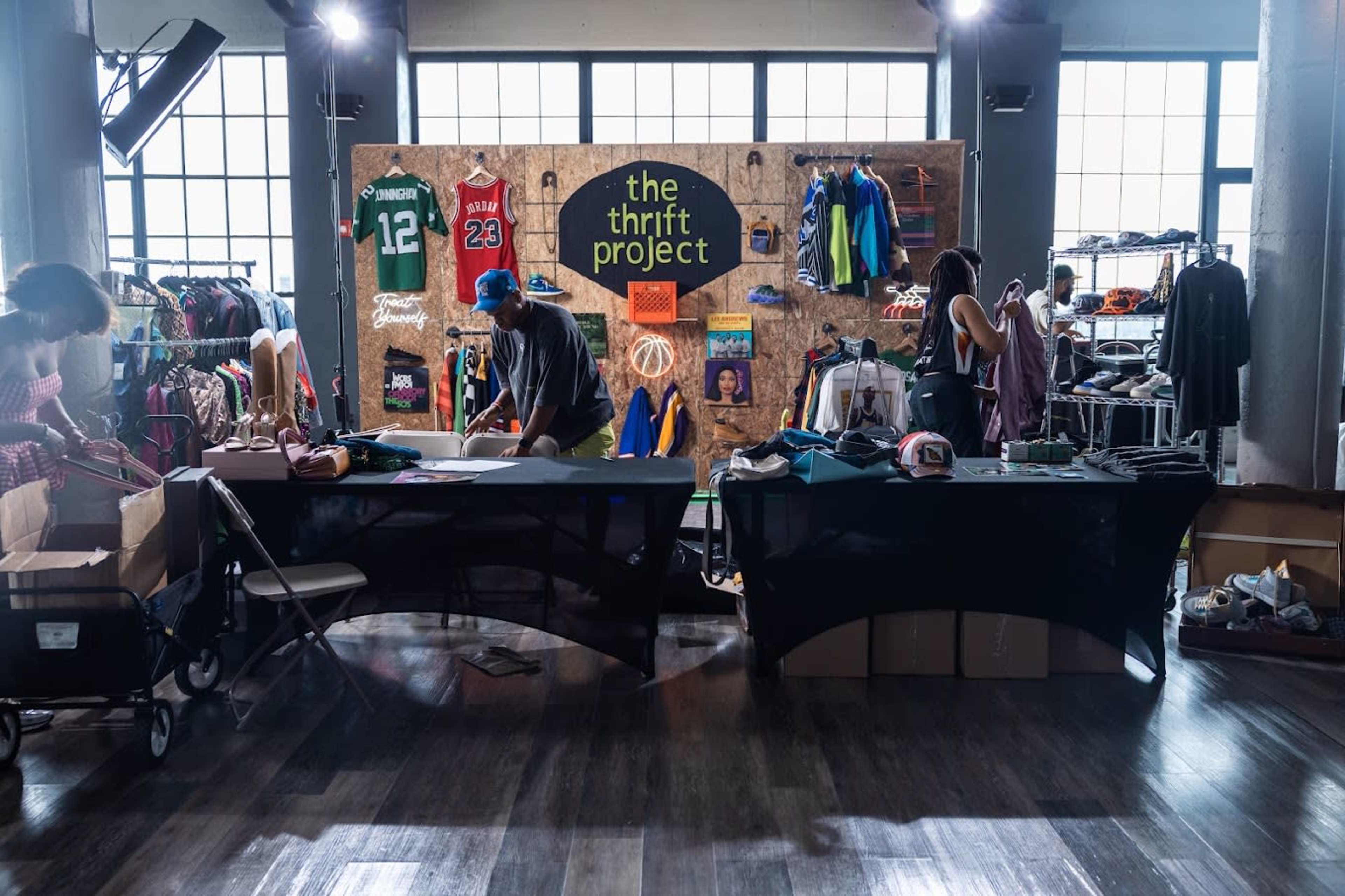 A thrift project event displays various clothing items on racks and tables, with people organizing and browsing through the merchandise.
