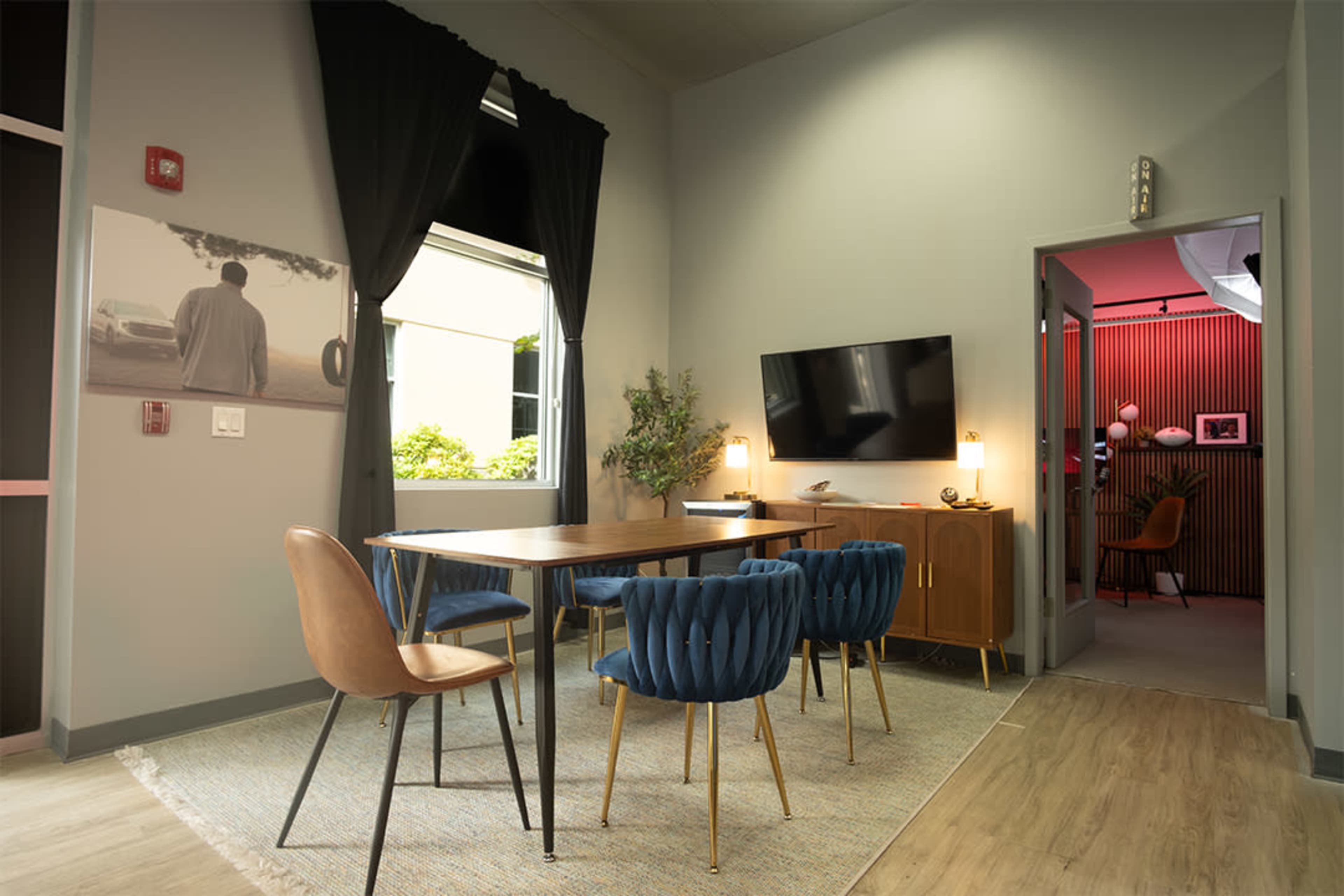 The image shows a modern conference room featuring a wooden table surrounded by blue and brown chairs, with a television on the wall and natural light coming through a window.