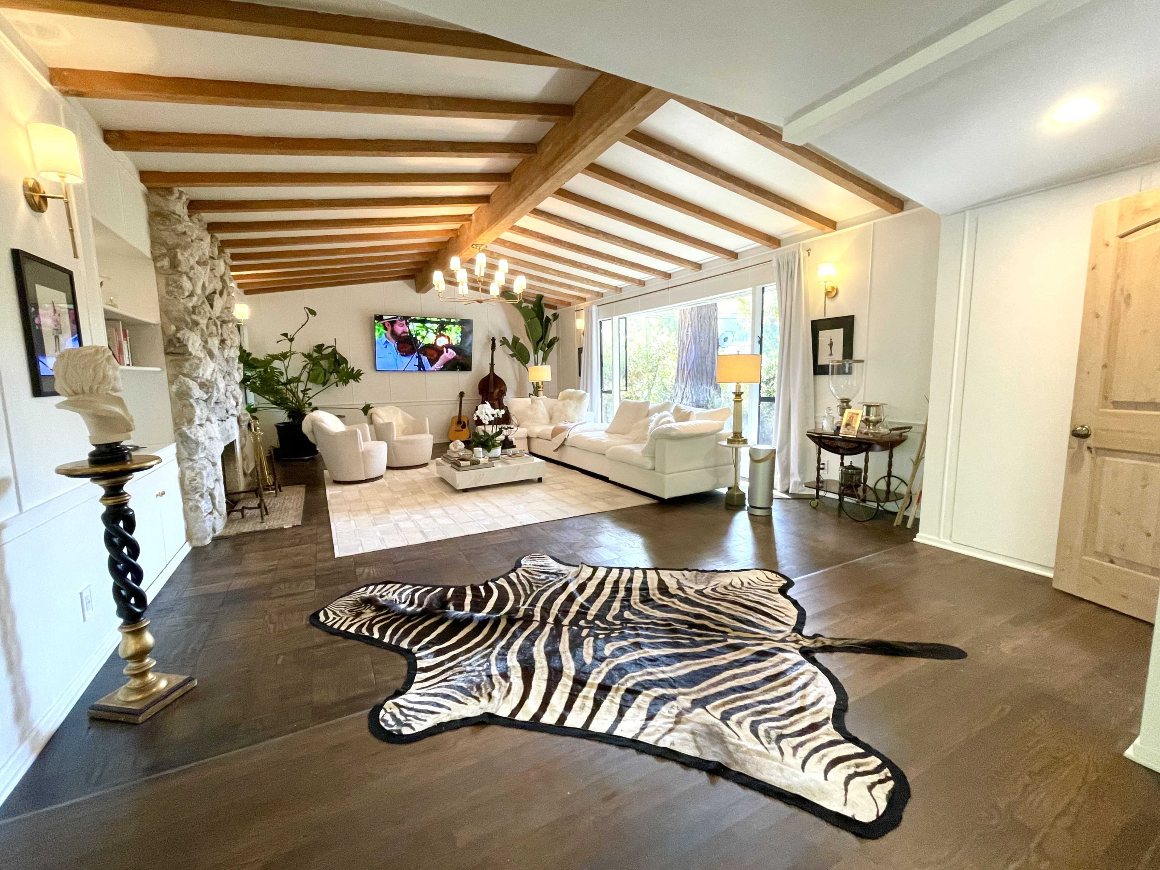 The image shows a spacious living room featuring a zebra-patterned rug, a stone accent wall, white furniture, and large windows allowing natural light to fill the space.